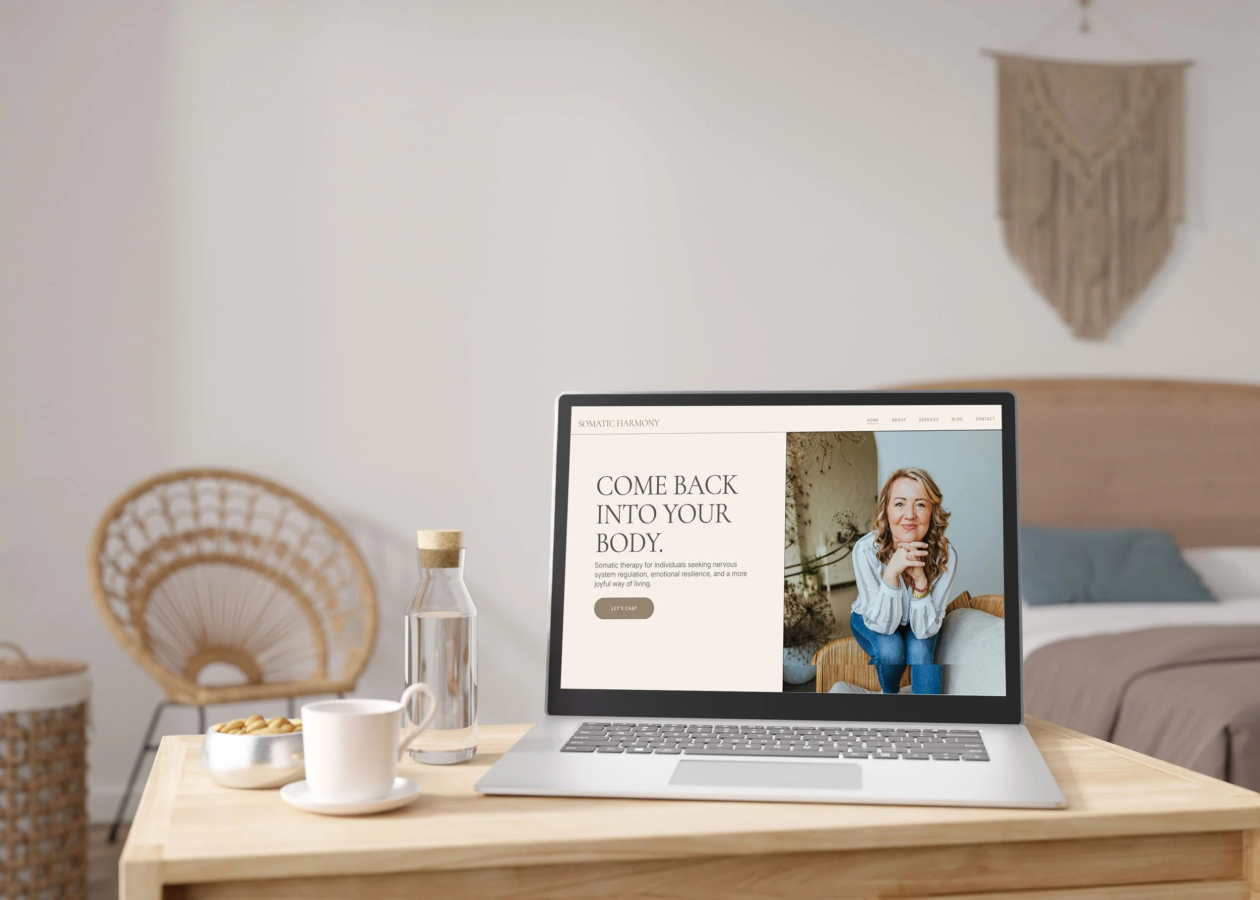 Laptop on a wooden desk displaying a website for somatic therapy, with a woman smiling in a photo on the screen. The desk has a glass of water, a cup, and a bowl of cookies. Background includes a wicker chair, a bed, and wall decorations in a cozy room.