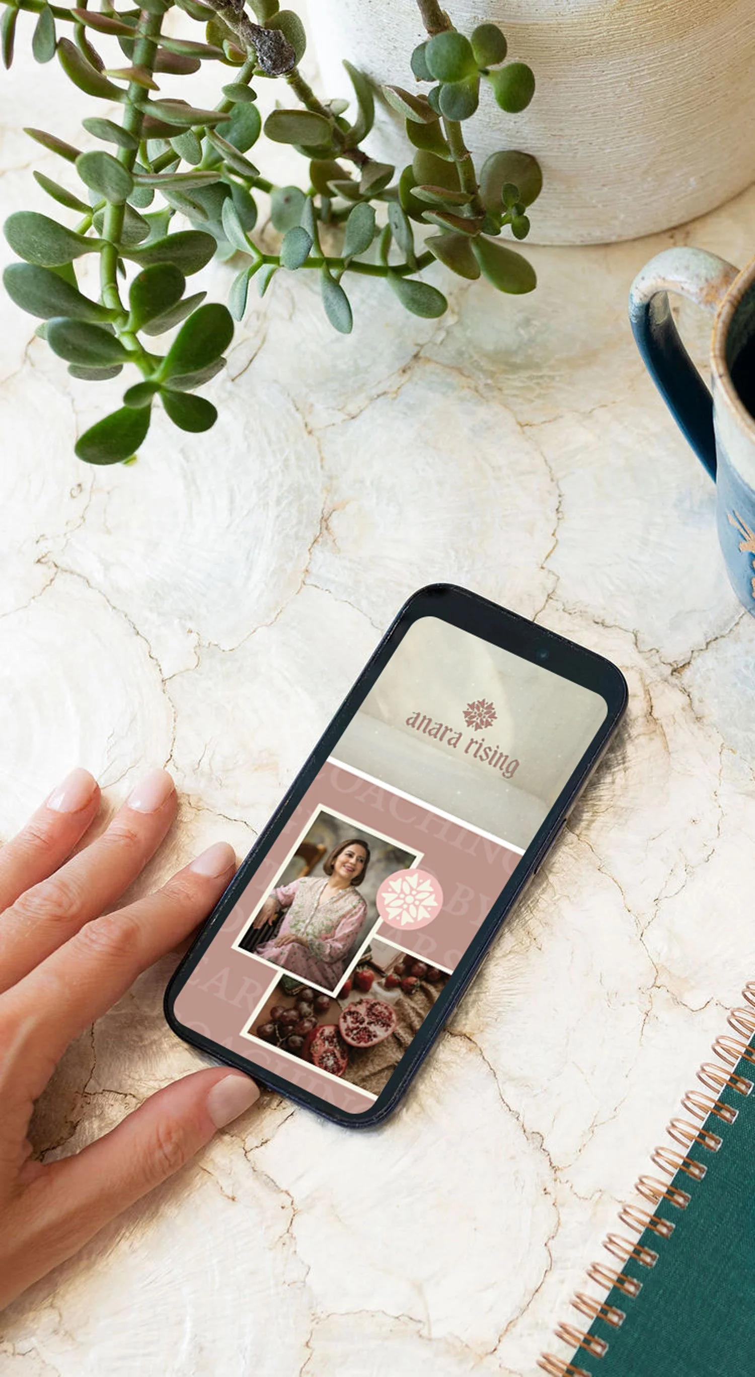 A hand holding a smartphone displaying a website or app called 'anara rising,' with a picture of a woman dressed in pink. The surface has a potted plant, a cup, a blue notebook, and a spiral notebook nearby.