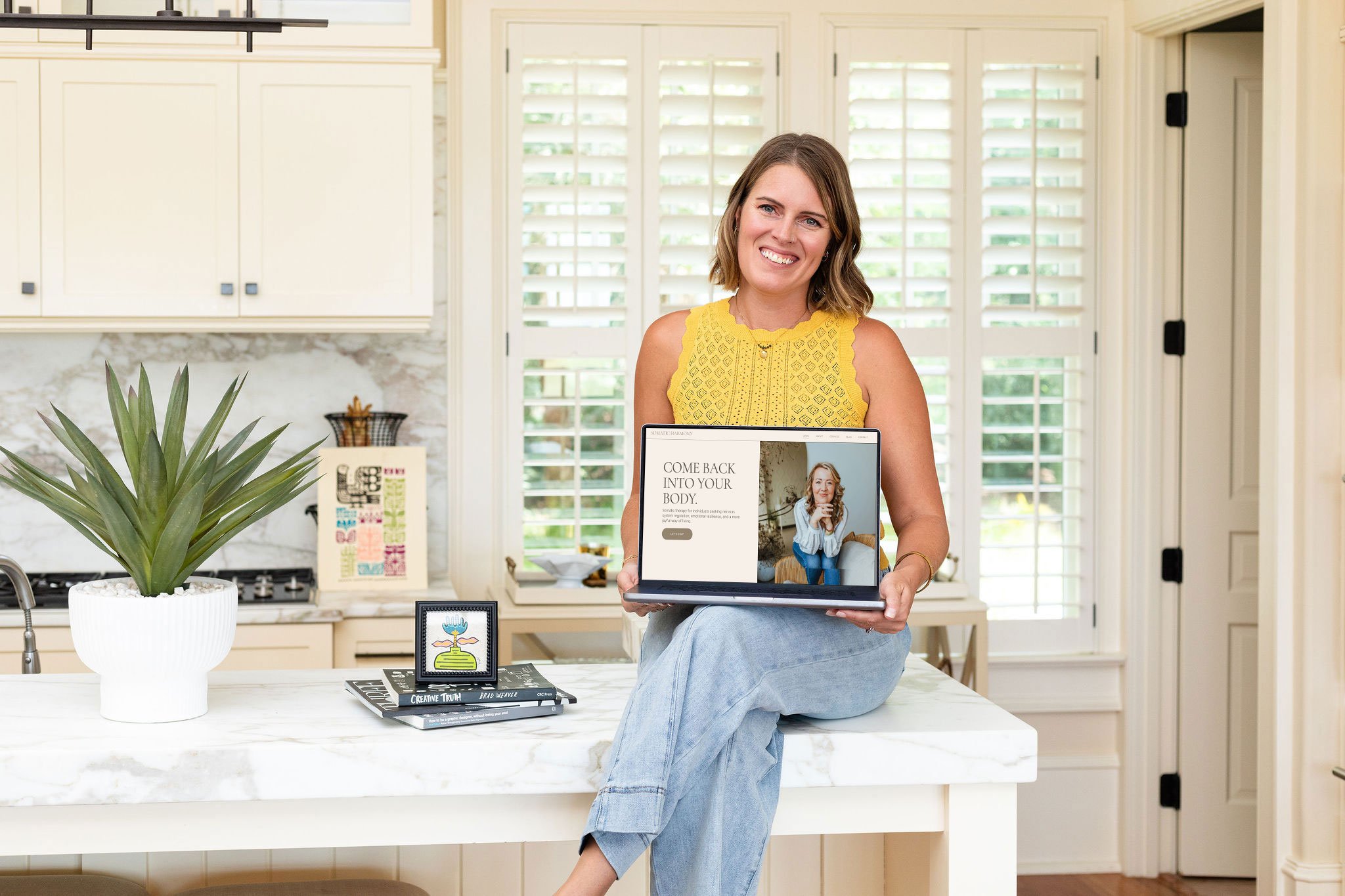 Woman sitting on kitchen counter holding a laptop with a website on the screen, next to a potted plant and various books and framed pictures.