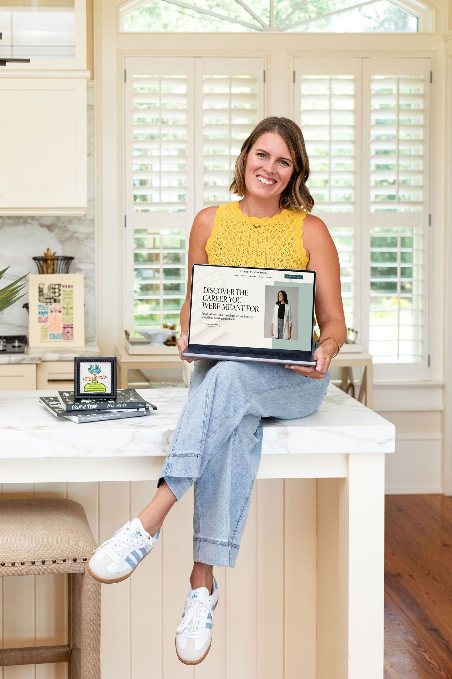 Woman sitting on kitchen counter holding her laptop, smiling at camera, wearing a yellow sleeveless top and light blue jeans, with a white and blue Adidas sneakers. Kitchen background with large windows with plantation shutters, books, and decorative items.