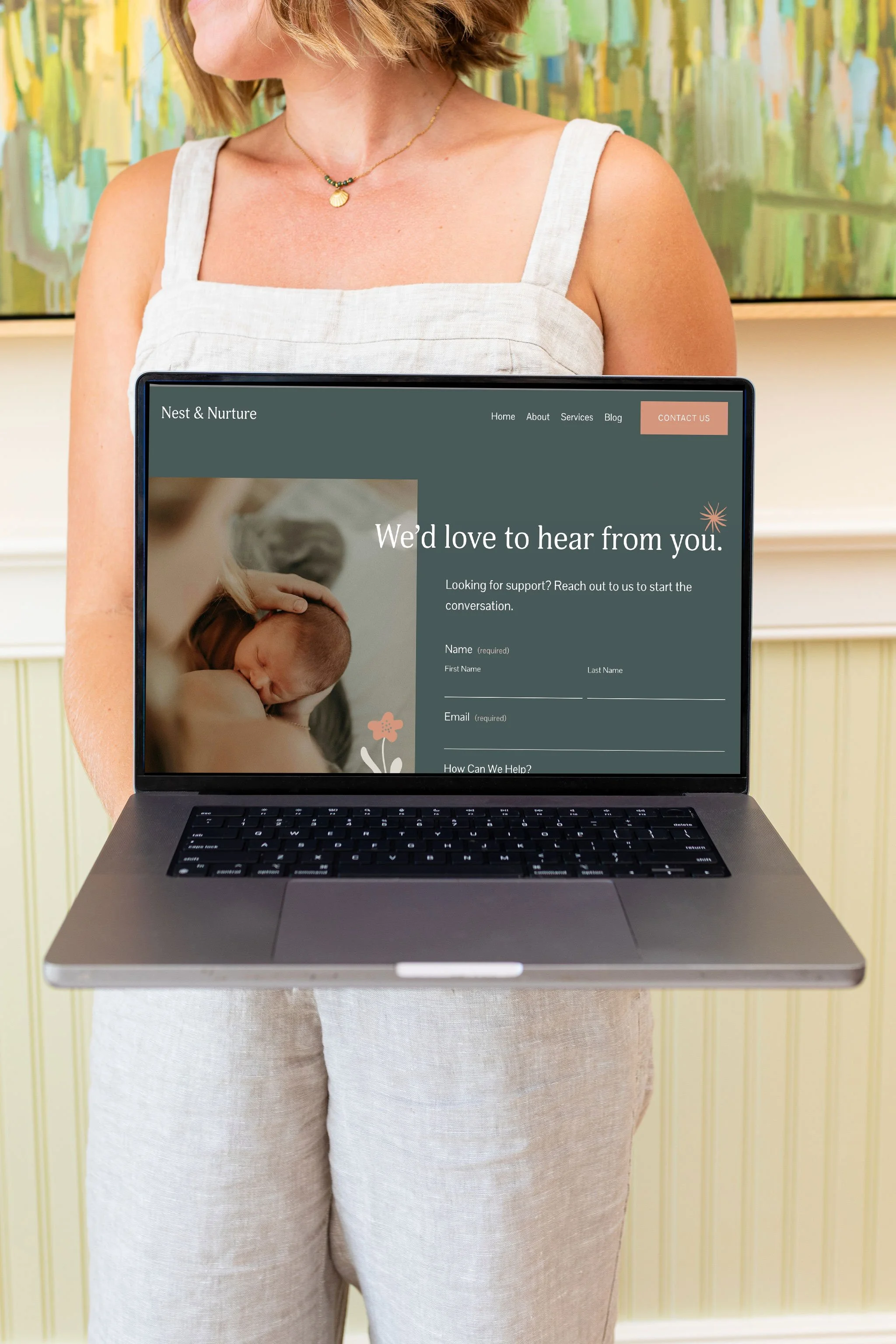 Rear view of a woman holding an open laptop displaying a website about nurturing and support, with her face partially visible, standing in front of a colorful abstract painting.