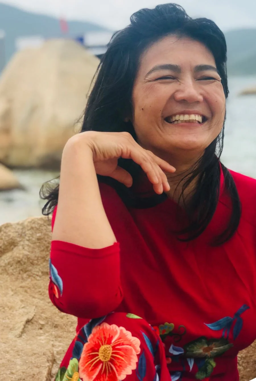 A woman smiling outdoors with her hand resting on her chin, wearing a red floral dress, with water and rocks in the background.