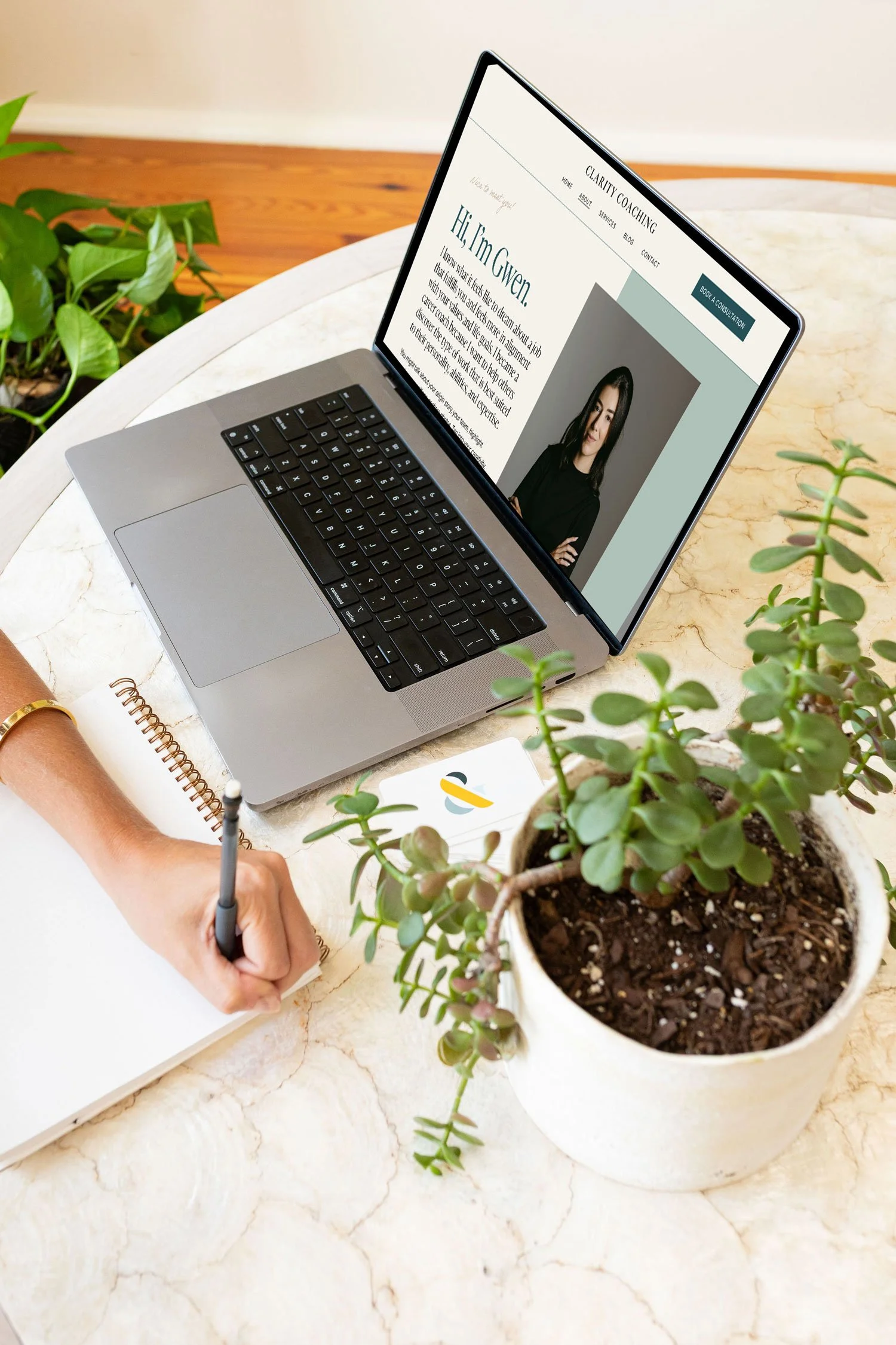 A person taking notes and working on a laptop at a marble table, with a potted plant and a notebook in the foreground.