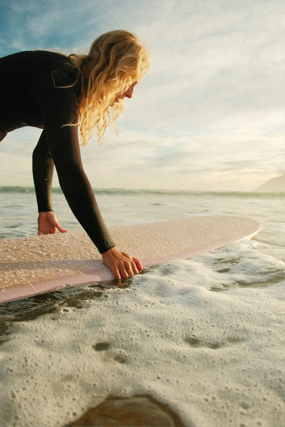A woman with blonde hair puts a surfboard into the ocean at the beach during sunset.