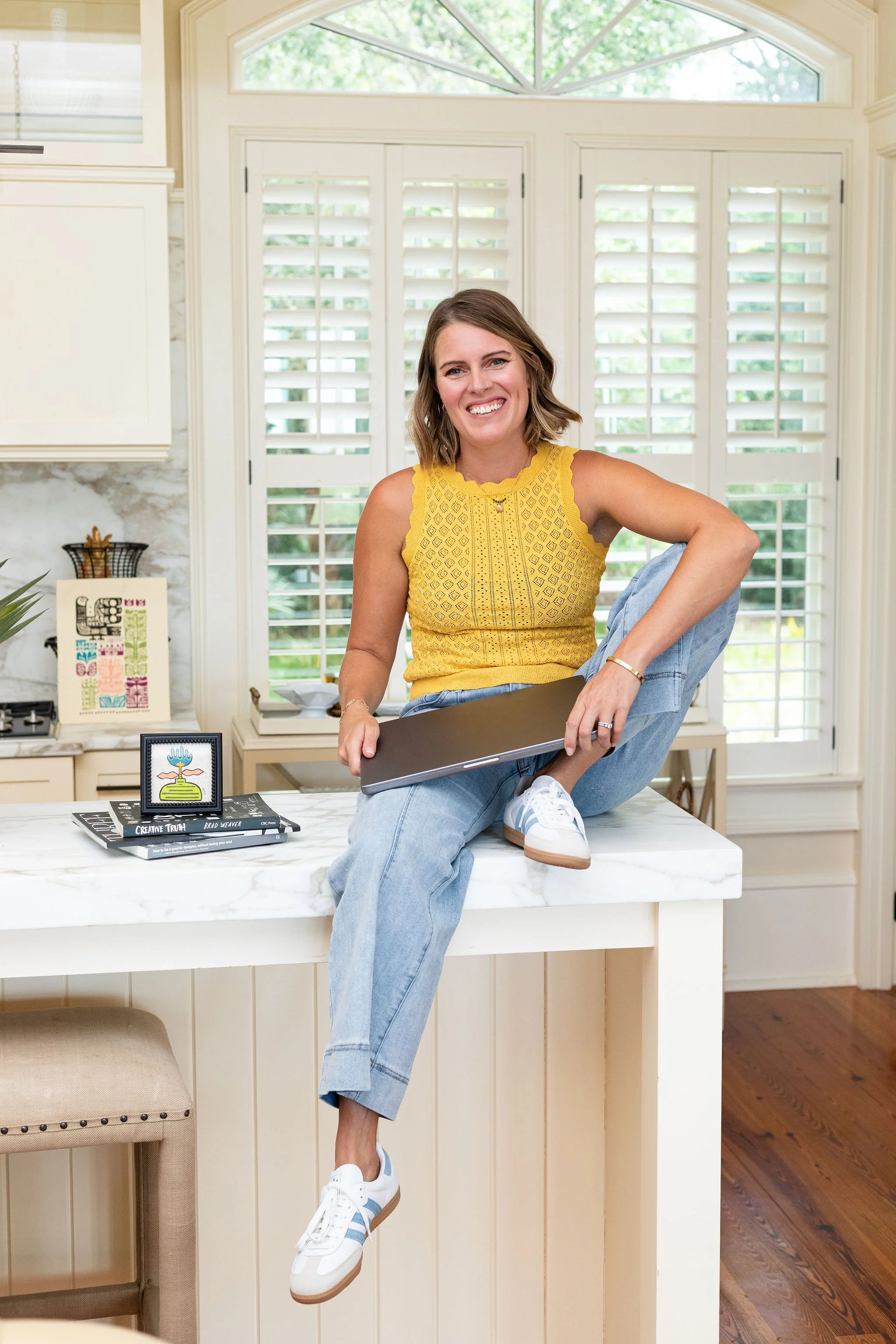 A woman with shoulder-length brown hair, wearing a yellow sleeveless top and light blue jeans, is sitting on a white kitchen island with her right leg crossed over her left knee. She is smiling and holding a closed laptop in her right hand. The background shows white shutters on windows, part of a kitchen with a marble countertop, some books and decorative items on the counter, and a wooden floor.