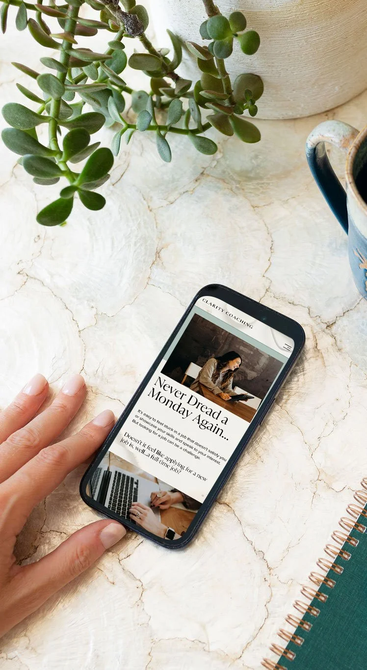 A person's hand holding a smartphone on a marble table, displaying a coaching website with the headline 'Never Dread Monday Again,' surrounded by a potted plant, a mug, and a notebook.
