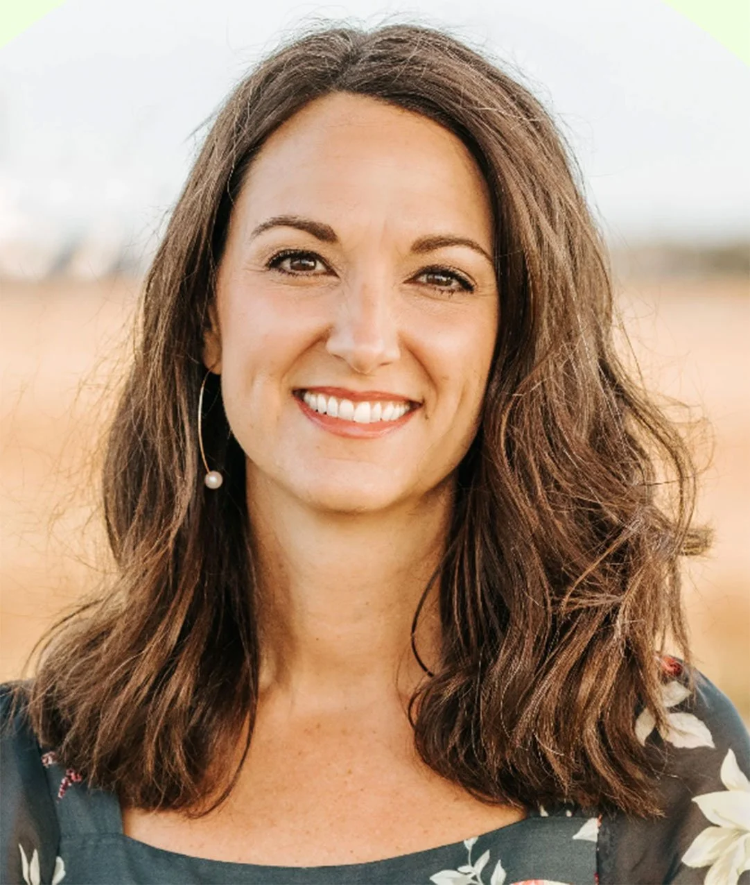 A woman with shoulder-length wavy brown hair, smiling, standing outdoors with a blurred natural background.