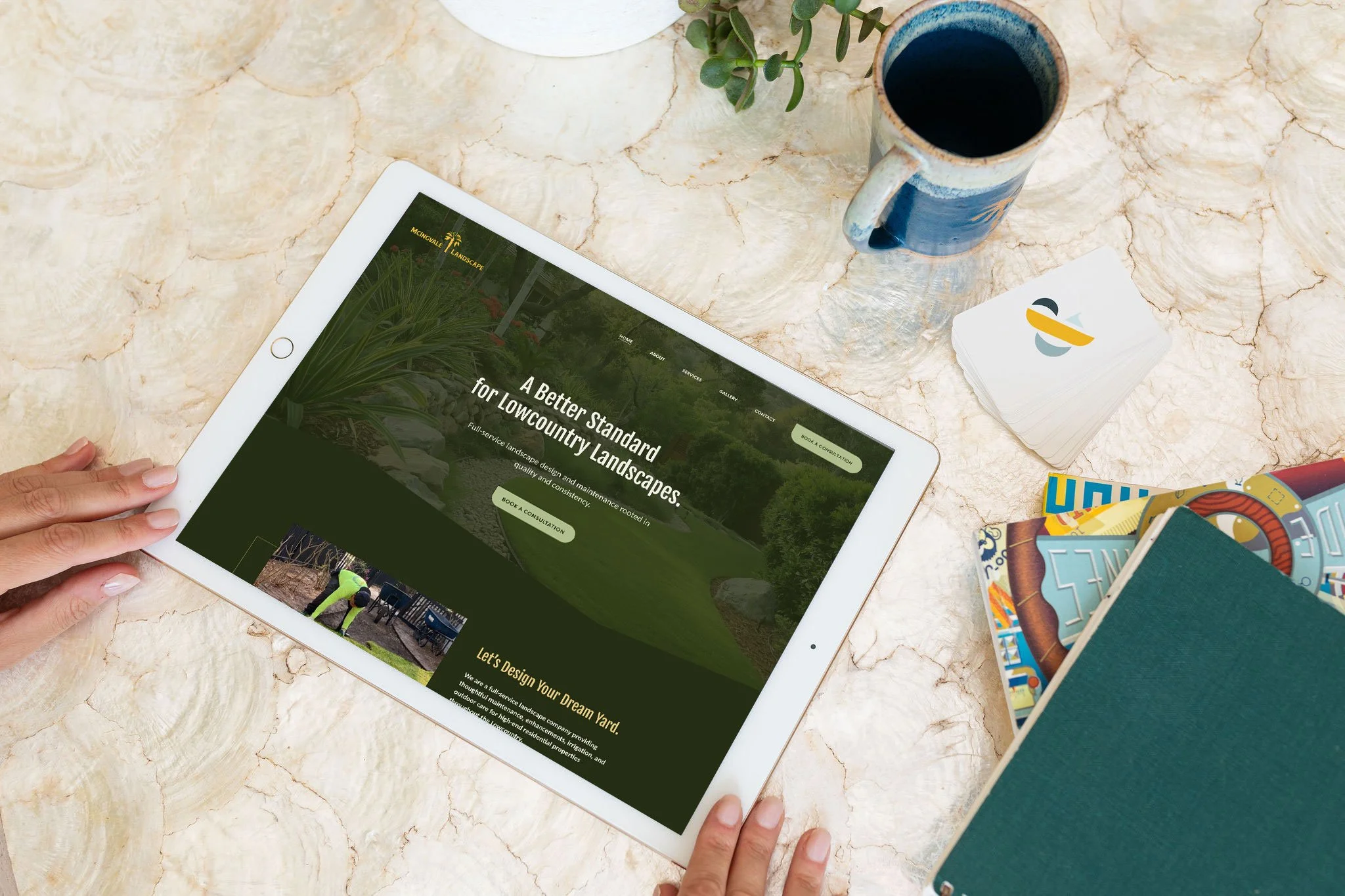 An electronic tablet displaying a landscaping company's website with the headline, 'A Better Standard for Lowcountry Landscapes.' The tablet rests on a marble surface, with a hand near the bottom left corner. Surrounding the tablet are a plant in a pot, a cup of dark coffee, a stack of business cards, and some colorful brochures or magazines.