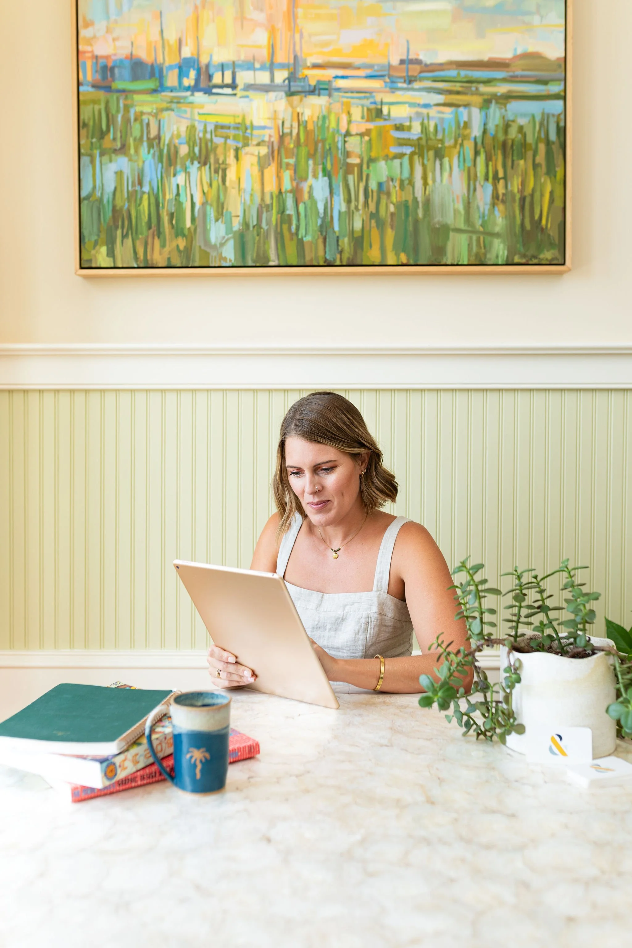 A woman with shoulder-length brown hair, wearing a sleeveless beige top and jewelry, sitting at a marble table, looking at a white digital tablet, with a potted plant, a blue and orange mug, books, and a closed notebook on the table, against a light-colored paneled wall and artwork hanging above.