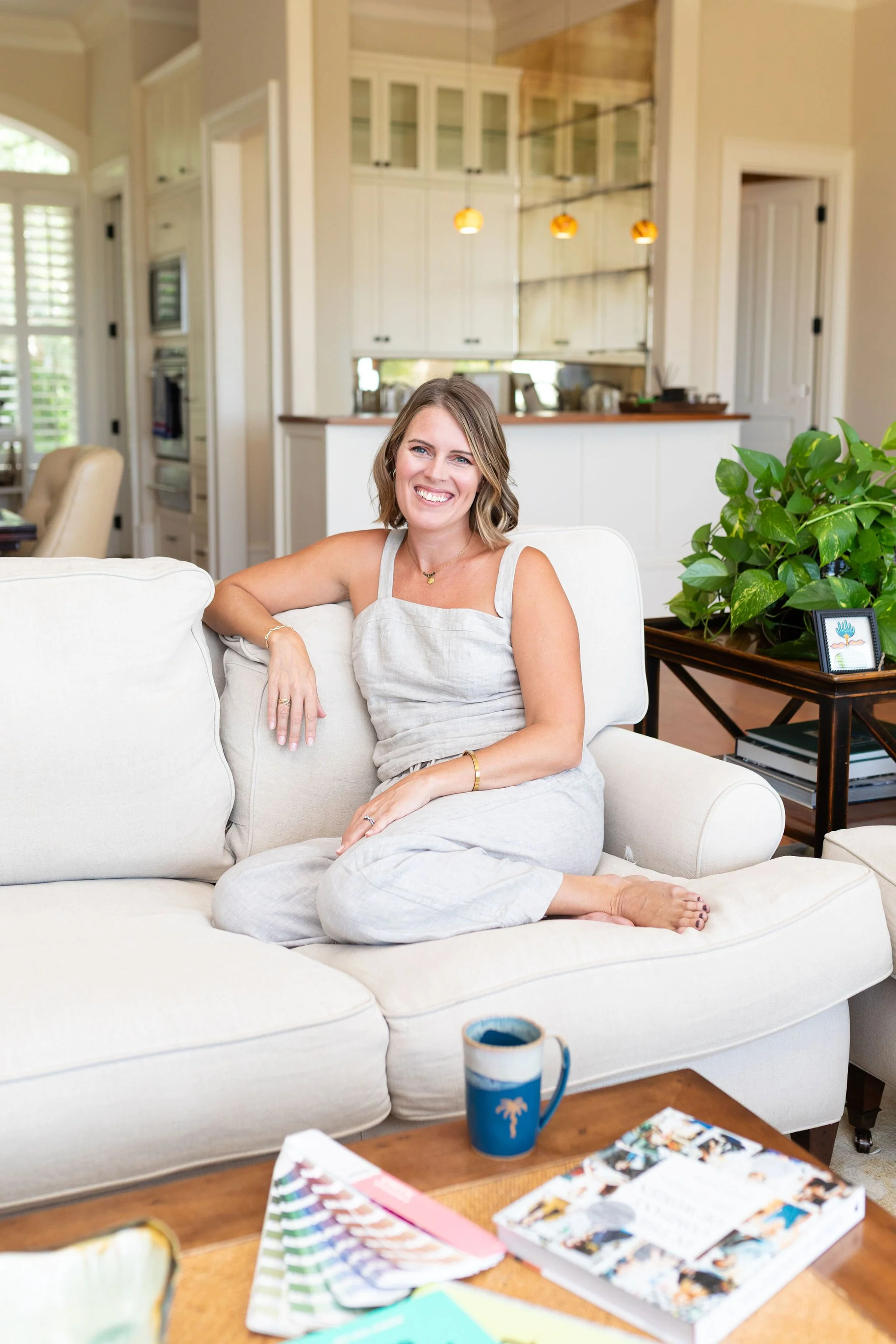 A woman with shoulder-length hair, wearing a light-colored dress, sitting happily on a white couch in a well-lit, modern kitchen and living room. There are colorful books and magazines on a wooden table in the foreground, and a large green potted plant with a framed picture nearby.