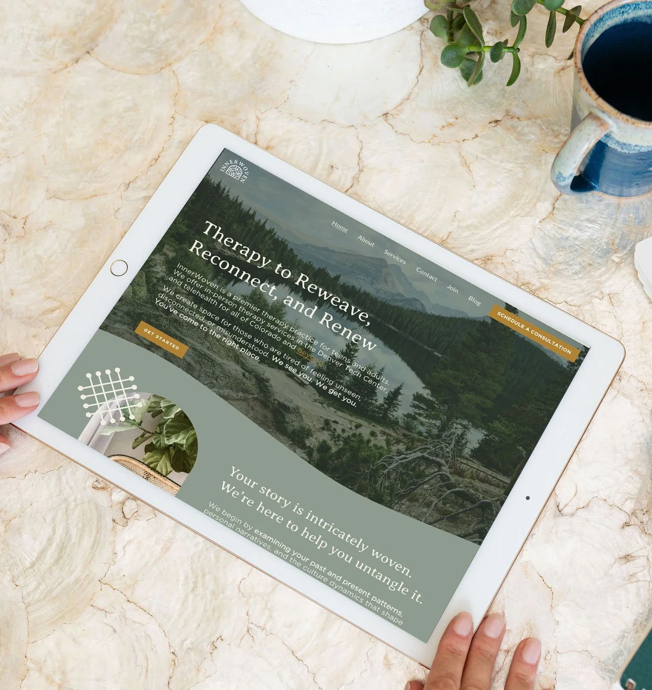 Tablet displaying website for InnerWoven, a therapy practice focused on reconnecting and renewing, with a scenic nature background, on a marble surface with a plant, mug, and white vase nearby.
