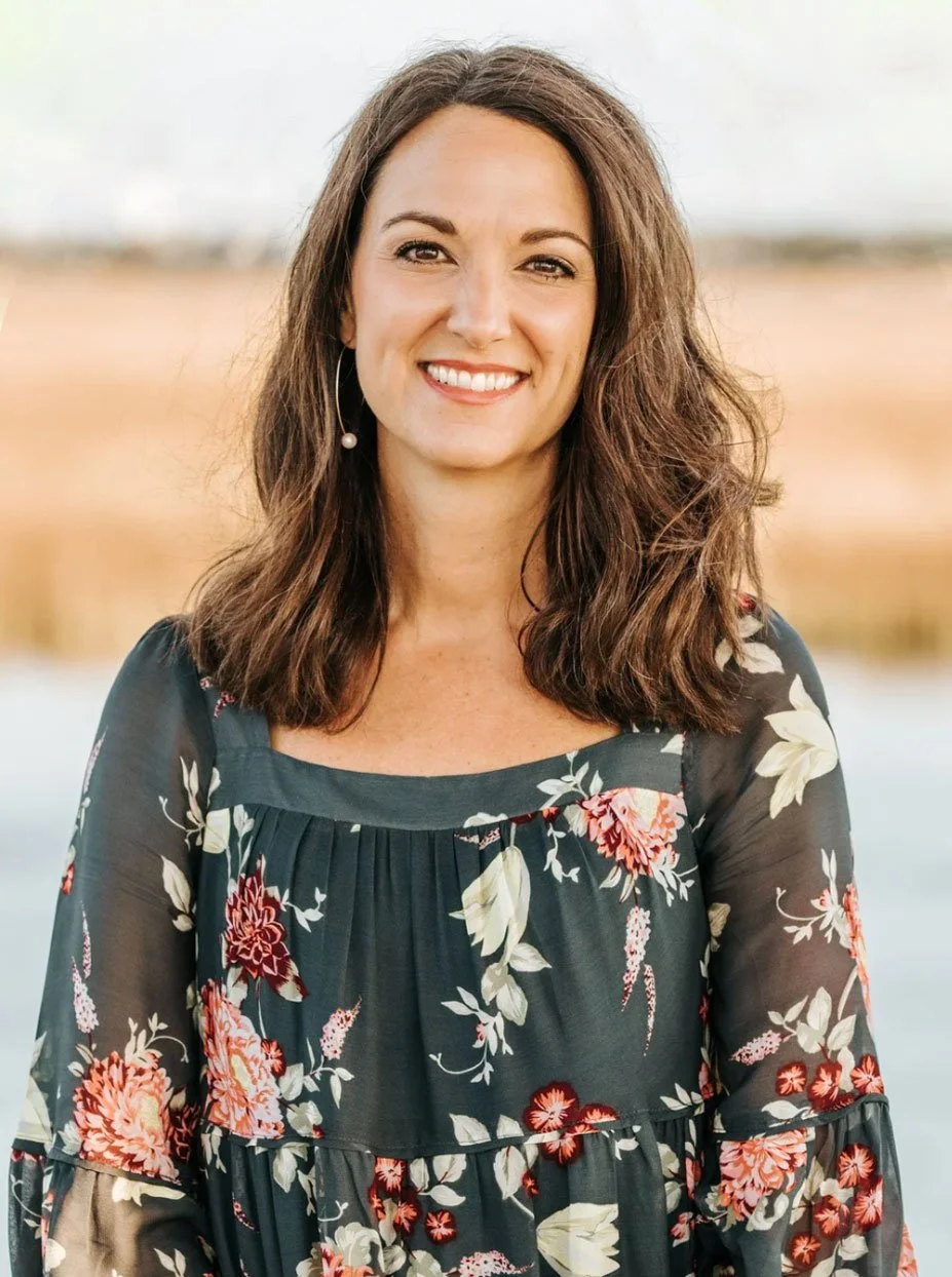 A woman with brown hair smiling outdoors near water, wearing a black floral dress and hoop earrings with a pearl pendant.