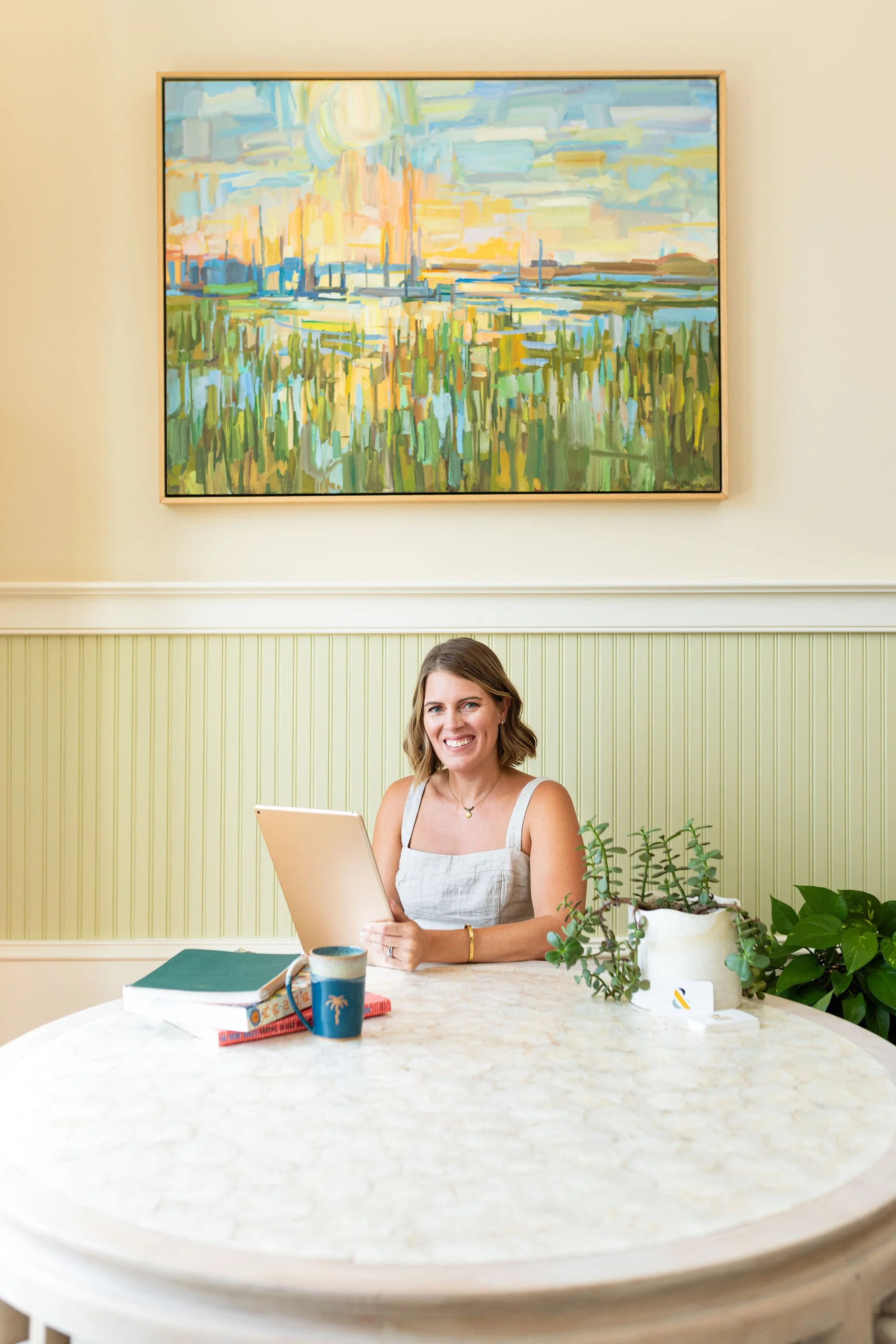 Callie Cullum, founder of The Look & The feel, sits at a table under a colorful painting, holding an iPad