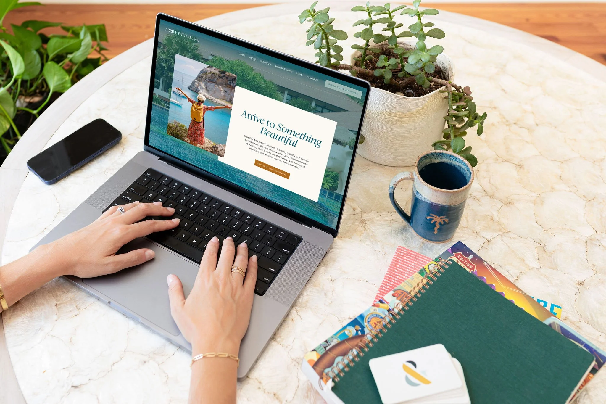 A person typing on a silver laptop on a stone table surrounded by green plants, notebooks, and a cup of coffee.