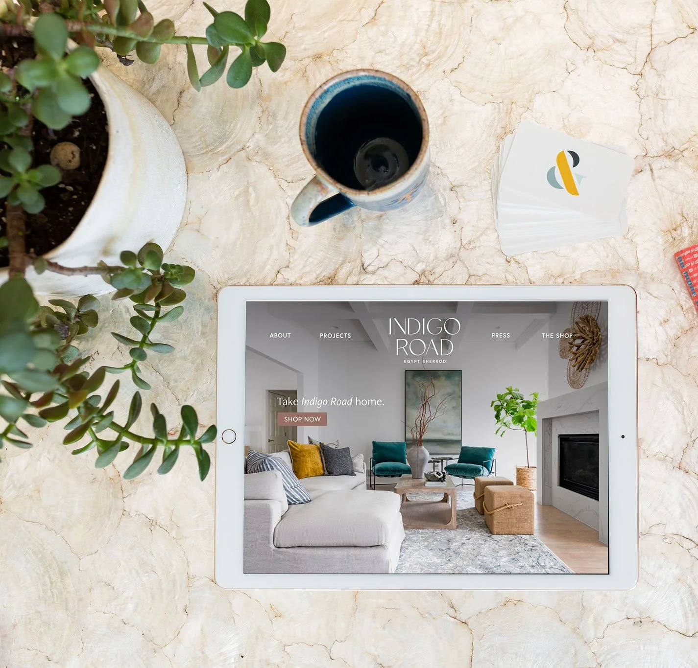 A tablet displaying a home interior website on a marble table. Next to the tablet are two potted plants, a black coffee cup, and a stack of white cards with a logo.