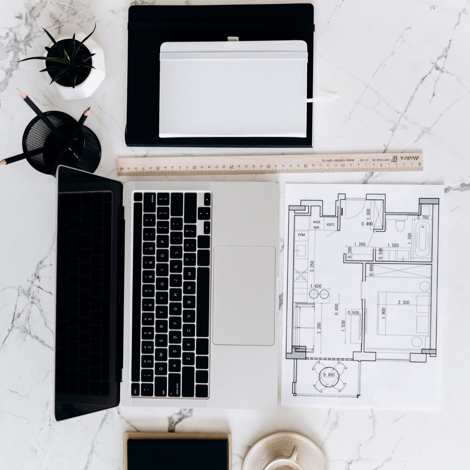 Desk with a laptop, blueprint, ruler, notebook, pens, potted plant, and smartphone on a white marble surface.