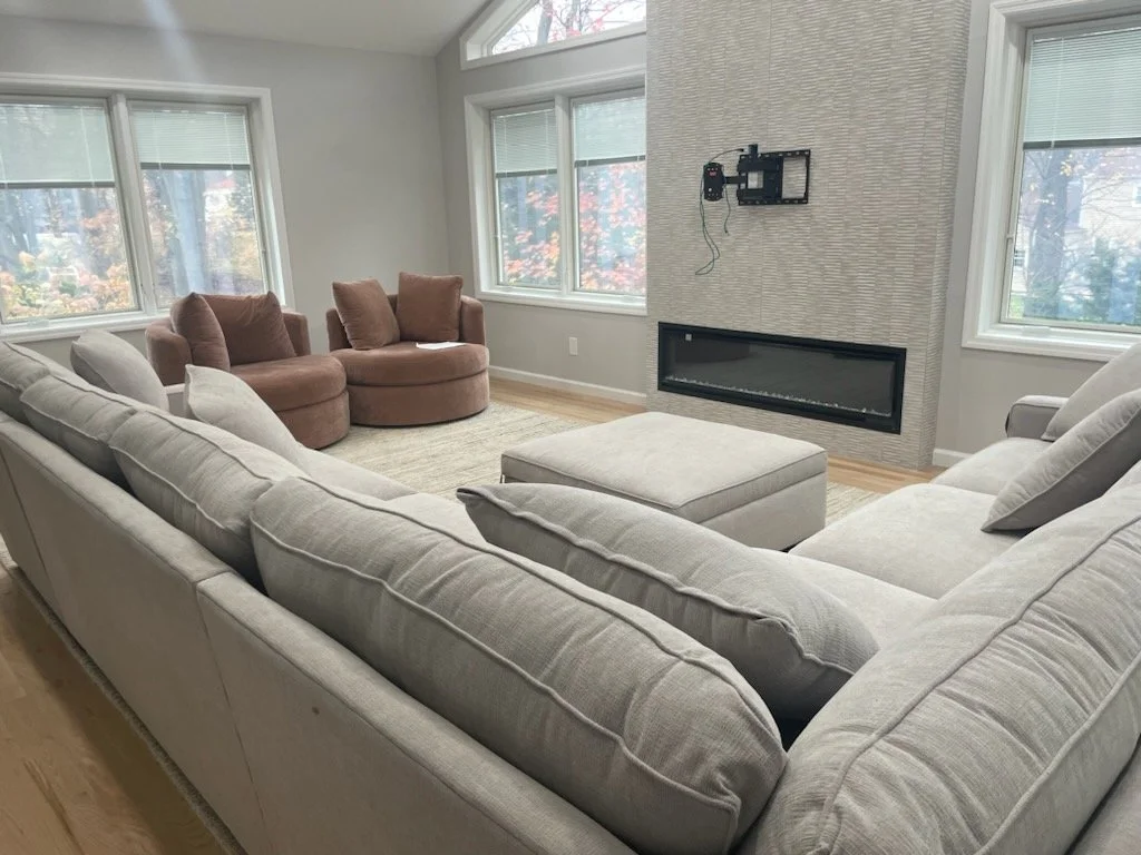 Living room with a large L-shaped white sofa, two pink armchairs, a beige ottoman, a wall-mounted fireplace, and multiple windows showing trees with autumn leaves.