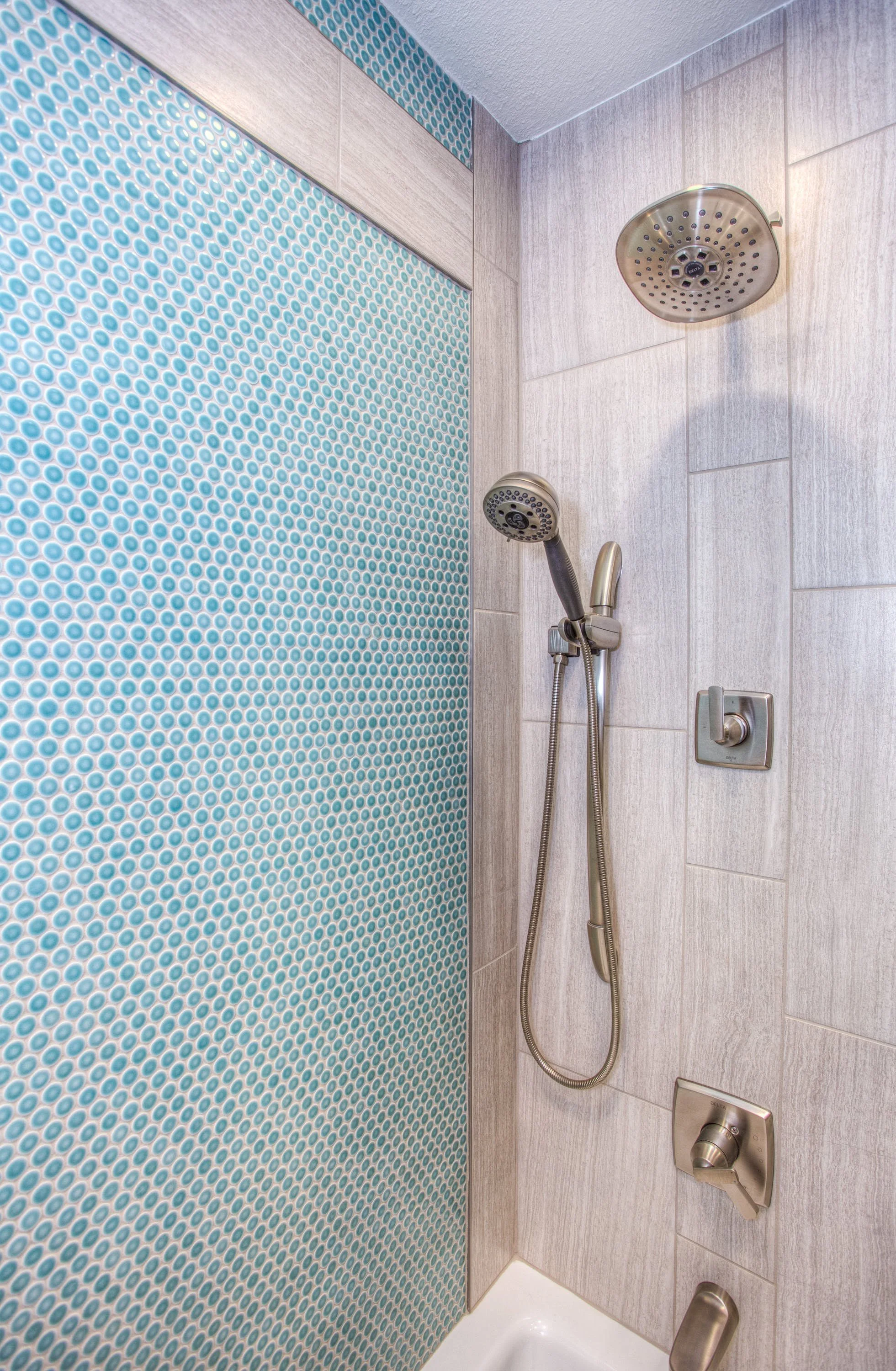 A shower area with light-colored tiled walls, a round overhead showerhead, a handheld showerhead attached by a flexible hose, a lever handle for water control, and a small white shower base.