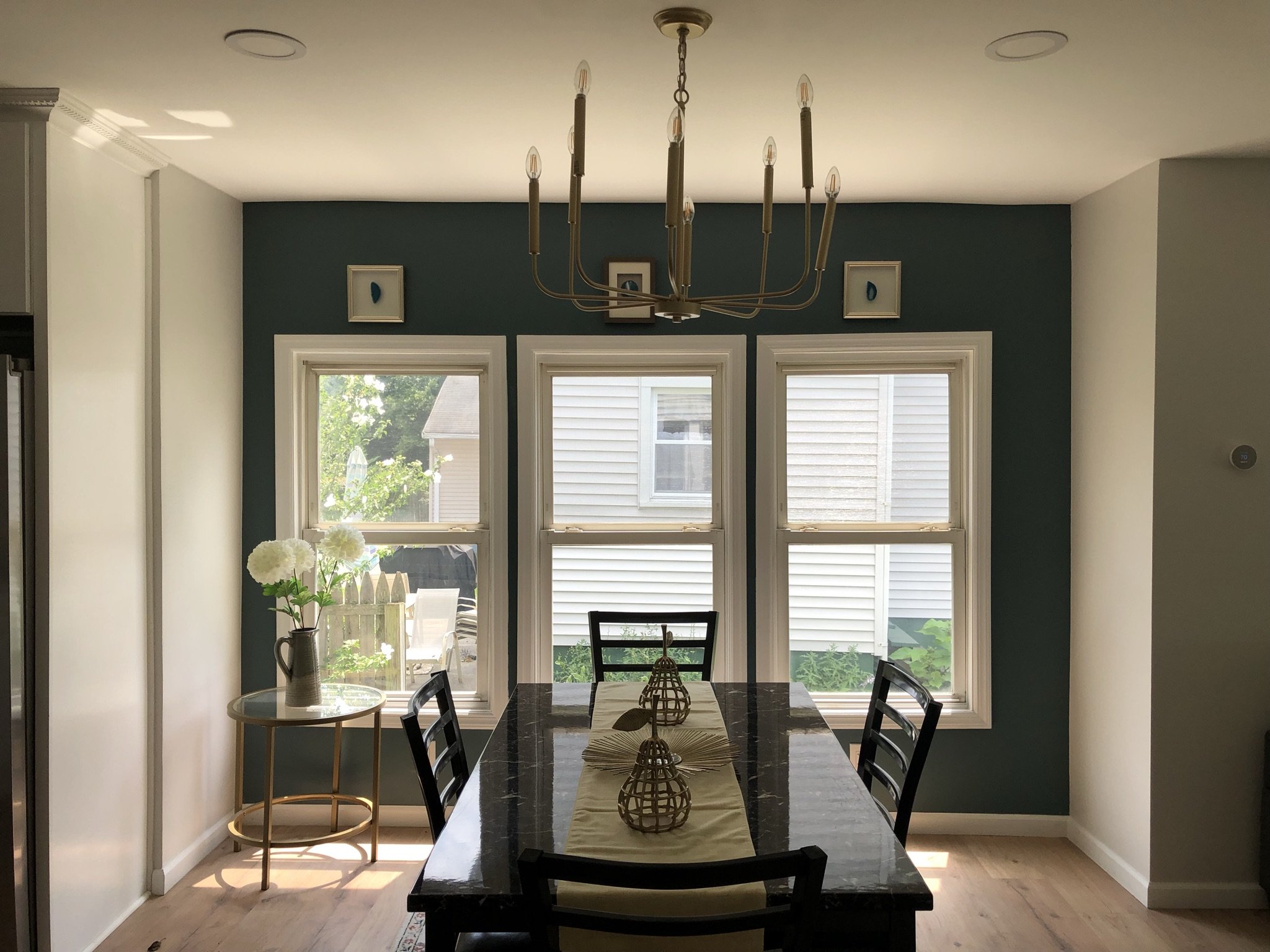 Dining room with a dark table, four chairs, a window wall, a chandelier, a side table with white flowers, and framed artwork.