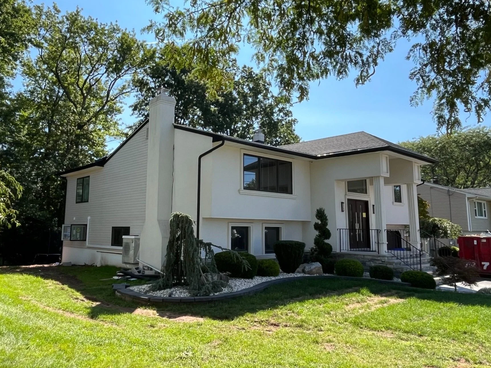 White two-story house with front steps, surrounded by a landscaped yard with bushes and a small tree, on a sunny day with trees in the background.