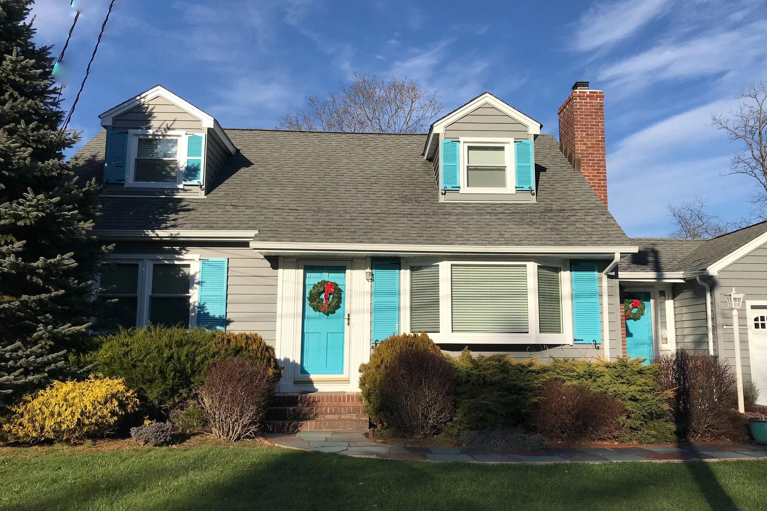 A two-story house decorated with Christmas wreaths on the doors, featuring blue shutters and a brick chimney, under a blue sky.