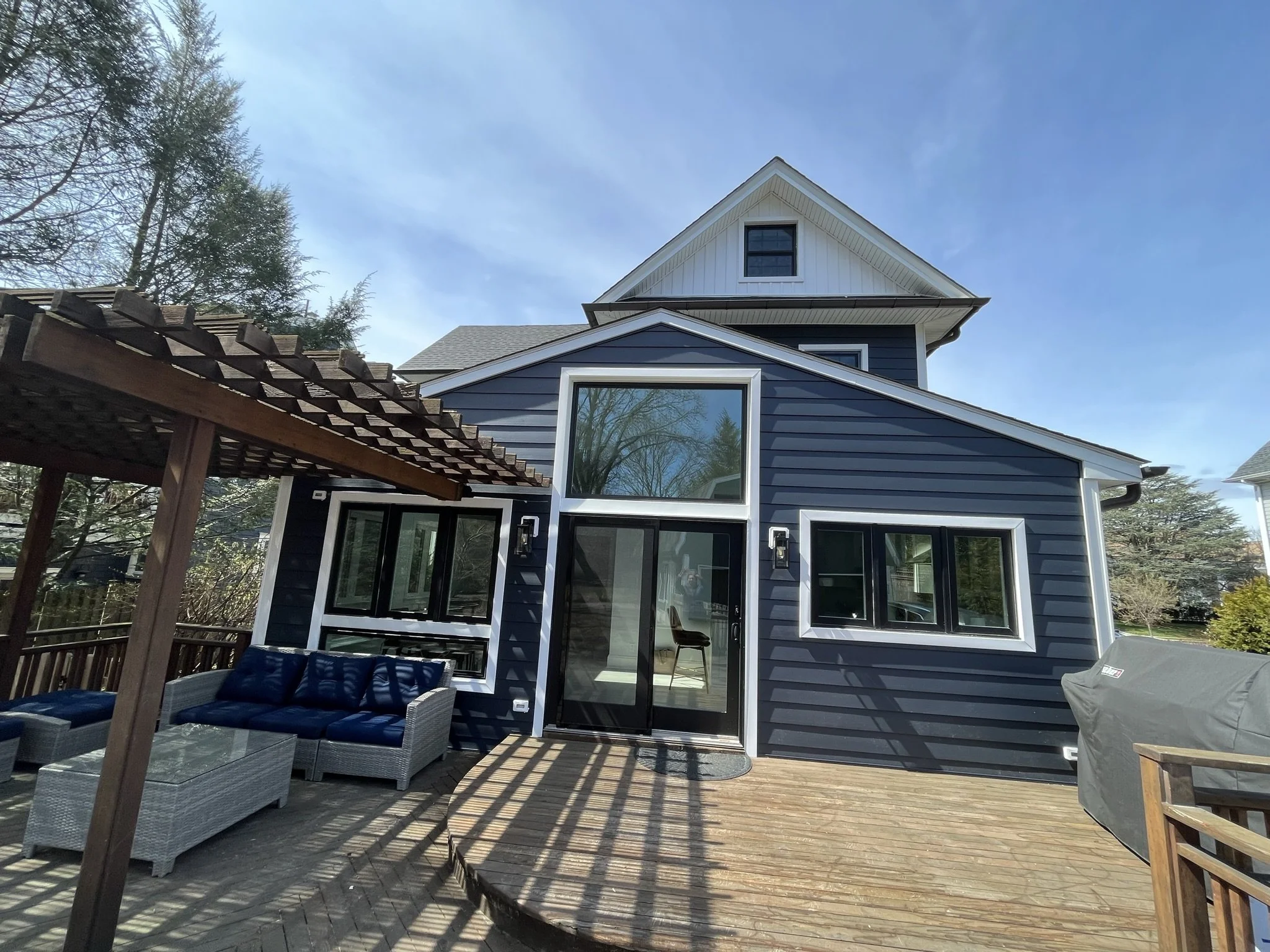 Backyard deck with outdoor furniture, a sliding glass door, and a house with blue siding and multiple windows under a partly cloudy sky.