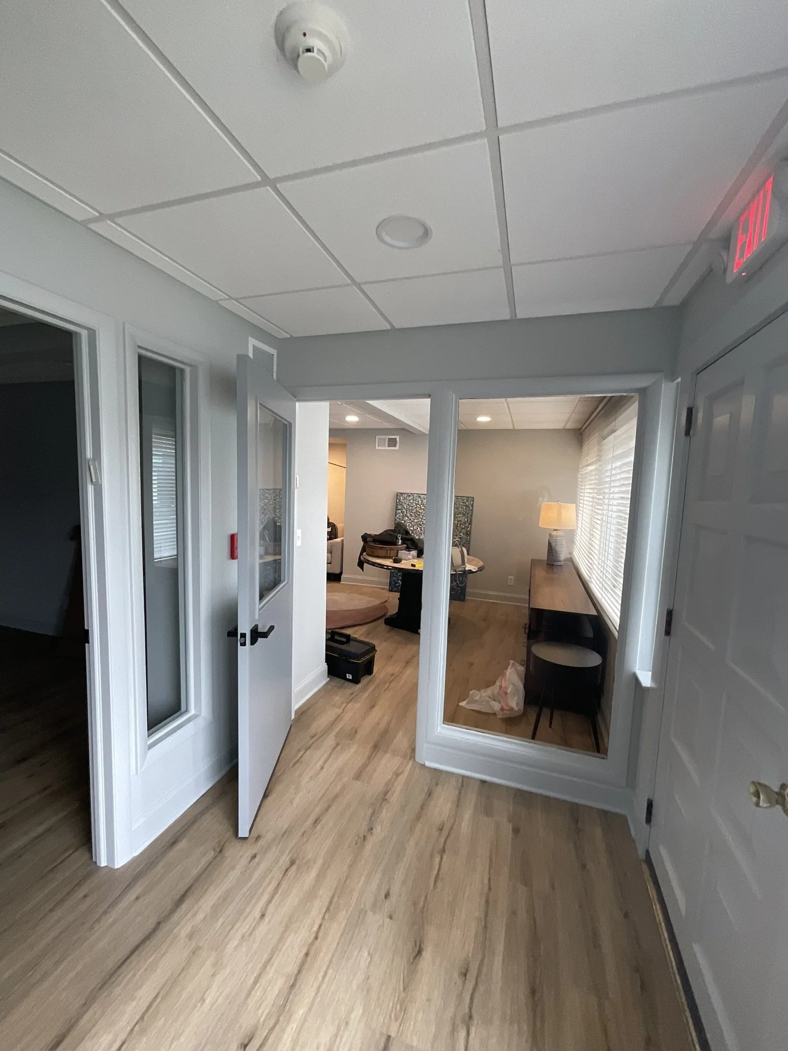 Interior view of a room with light-colored wooden flooring, a white door with glass panel open, a glass wall, a small wooden desk with a chair, and a window with white blinds. There is a table lamp, some bags, and a box inside.