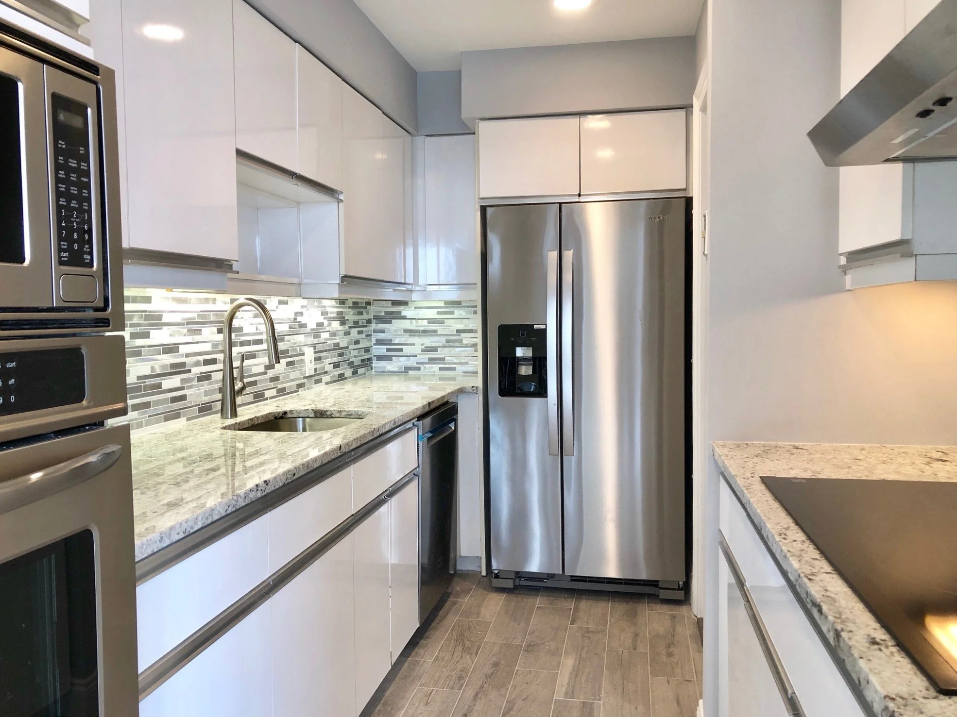 Modern kitchen with white cabinets, stainless steel refrigerator with water and ice dispenser, granite countertops, mosaic tile backsplash, microwave oven, electric cooktop, and wooden flooring.