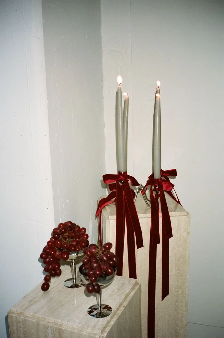 Two lit candles tied with red ribbons, with another bunch of grapes in two wine glasses.
