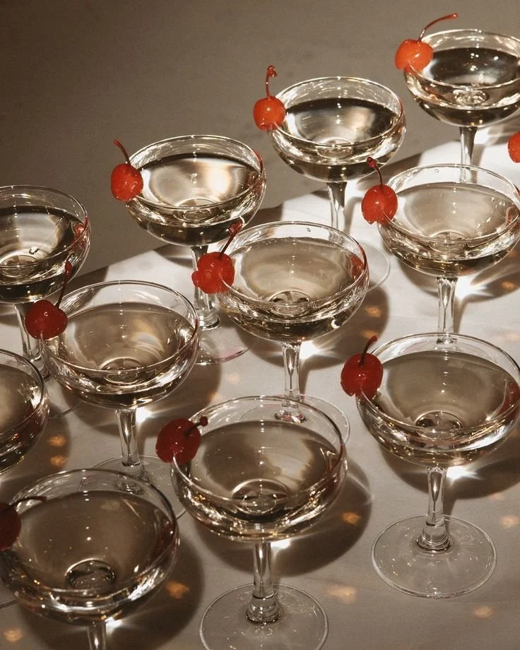 Multiple coupe glasses filled with a clear beverage, each garnished with a maraschino cherry on the rim, arranged on a white surface.