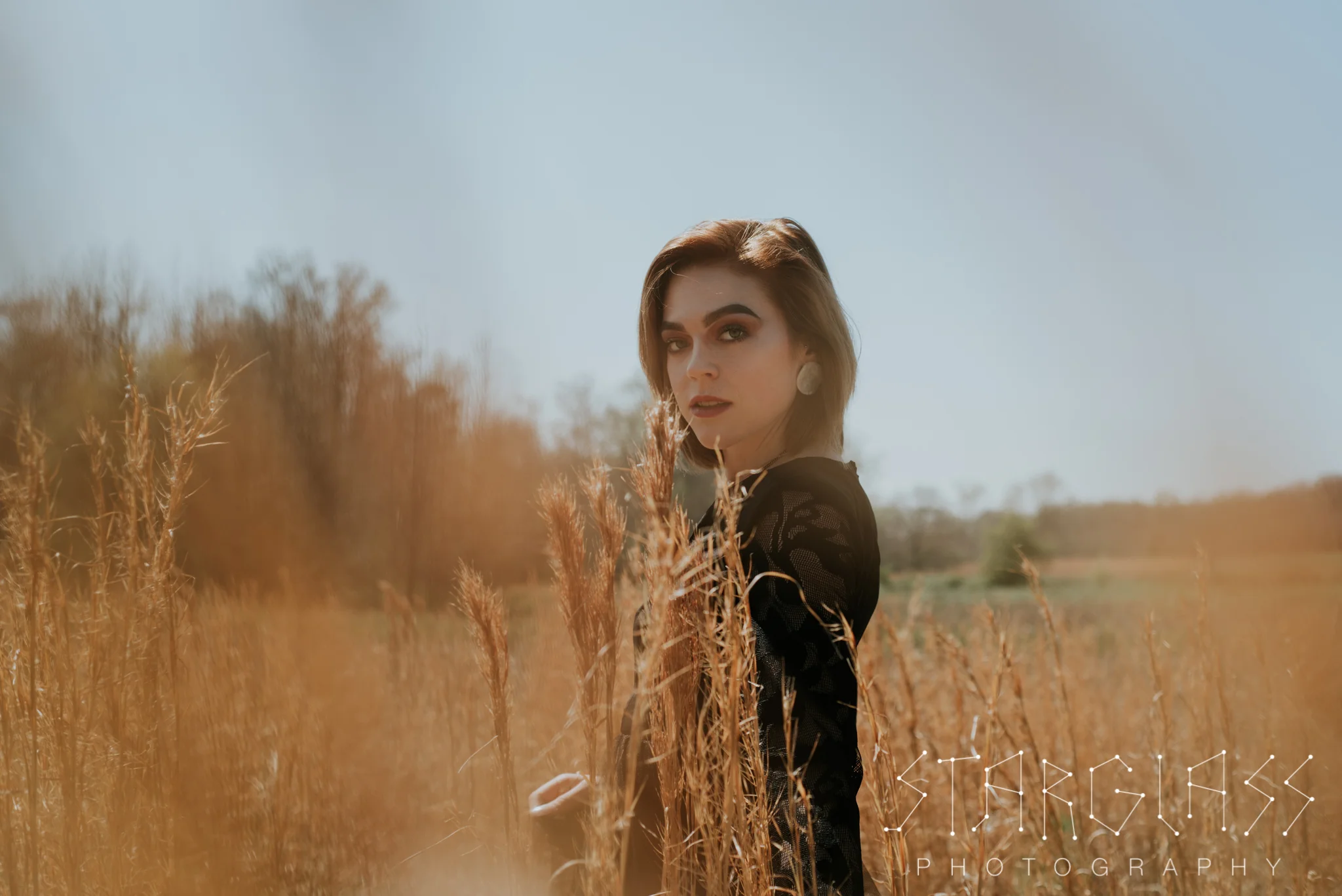Woman in a black dress standing in a field of tall golden grass under a cloudless blue sky.