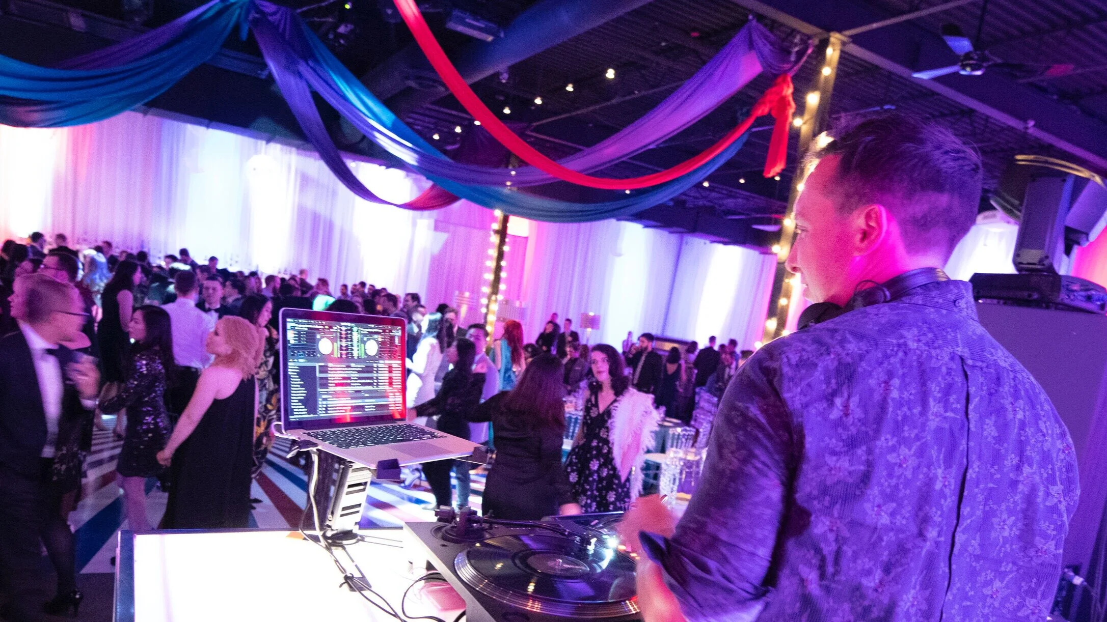 A DJ man with headphones standing at a mixer and laptop in a crowded dance hall decorated with drapes and string lights, people dancing and socializing.