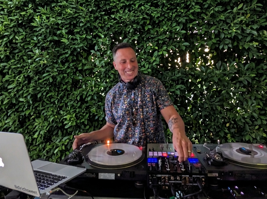 A DJ is smiling and adjusting music equipment outdoors with green shrubbery in the background.