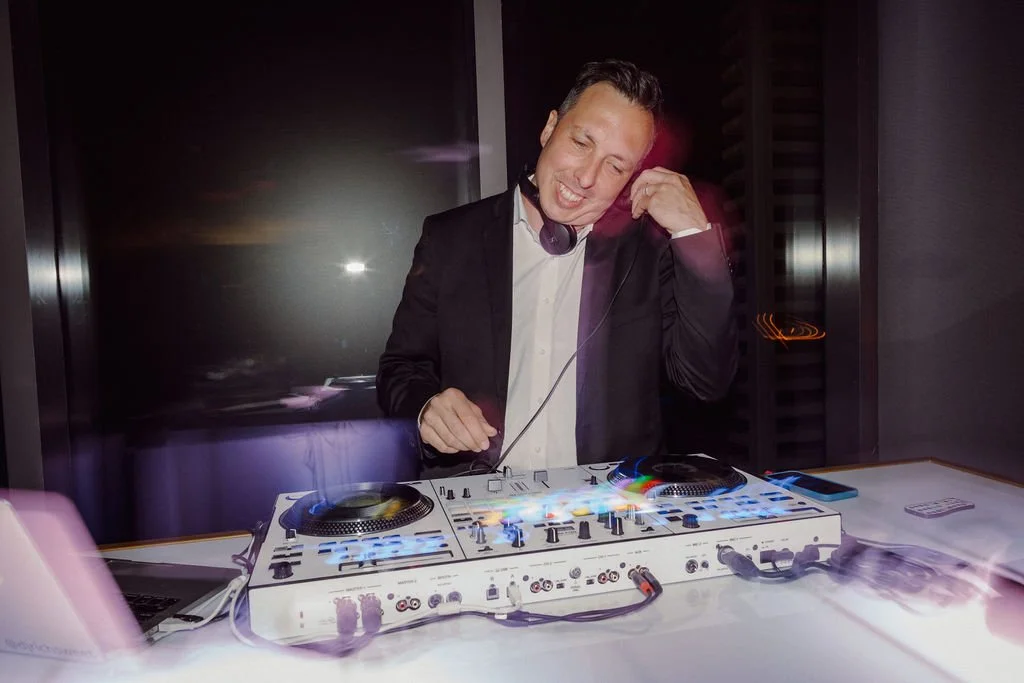 A man in a suit is DJing with headphones, smiling, at a DJ setup with a laptop and turntables in a dimly lit room.