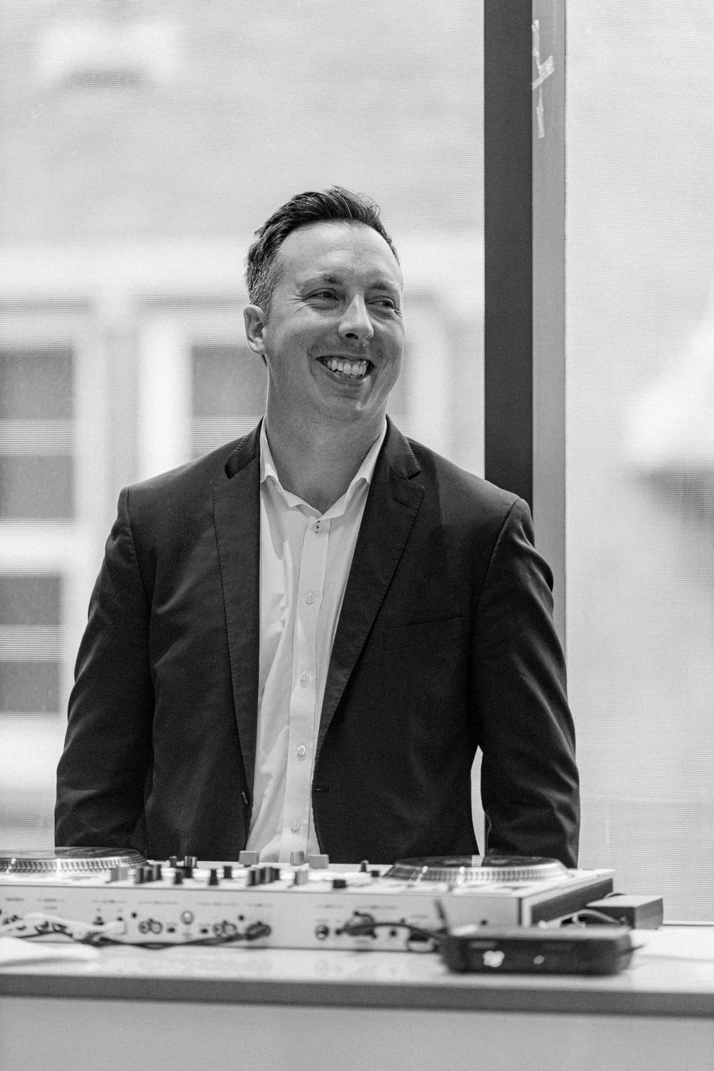 A man in a suit and tie smiling, standing behind DJ equipment in an indoor setting.