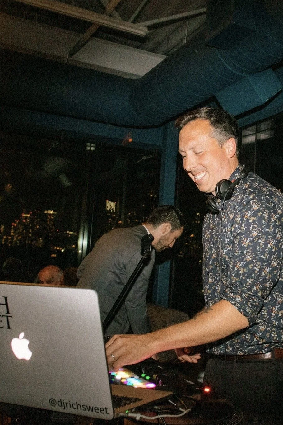 Two men at a nighttime event with a city skyline view in the background. One man is smiling, wearing a floral patterned shirt, and has headphones around his neck. The other man is bowing, dressed in a suit. There is a DJ equipment setup with a laptop that has a sticker reading '@dirichsweet'.