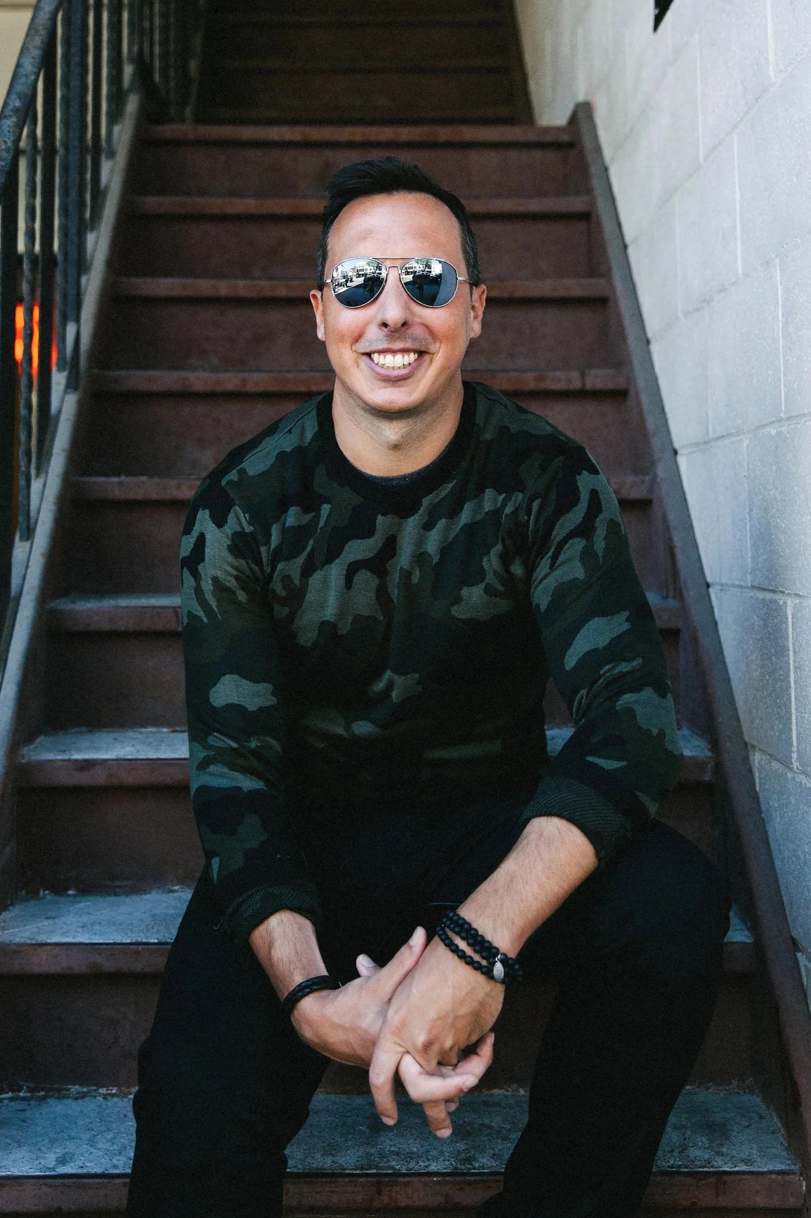 A man smiling and wearing reflective sunglasses, a camouflage long-sleeve shirt, and black pants, sitting on outdoor stairs near a white wall.