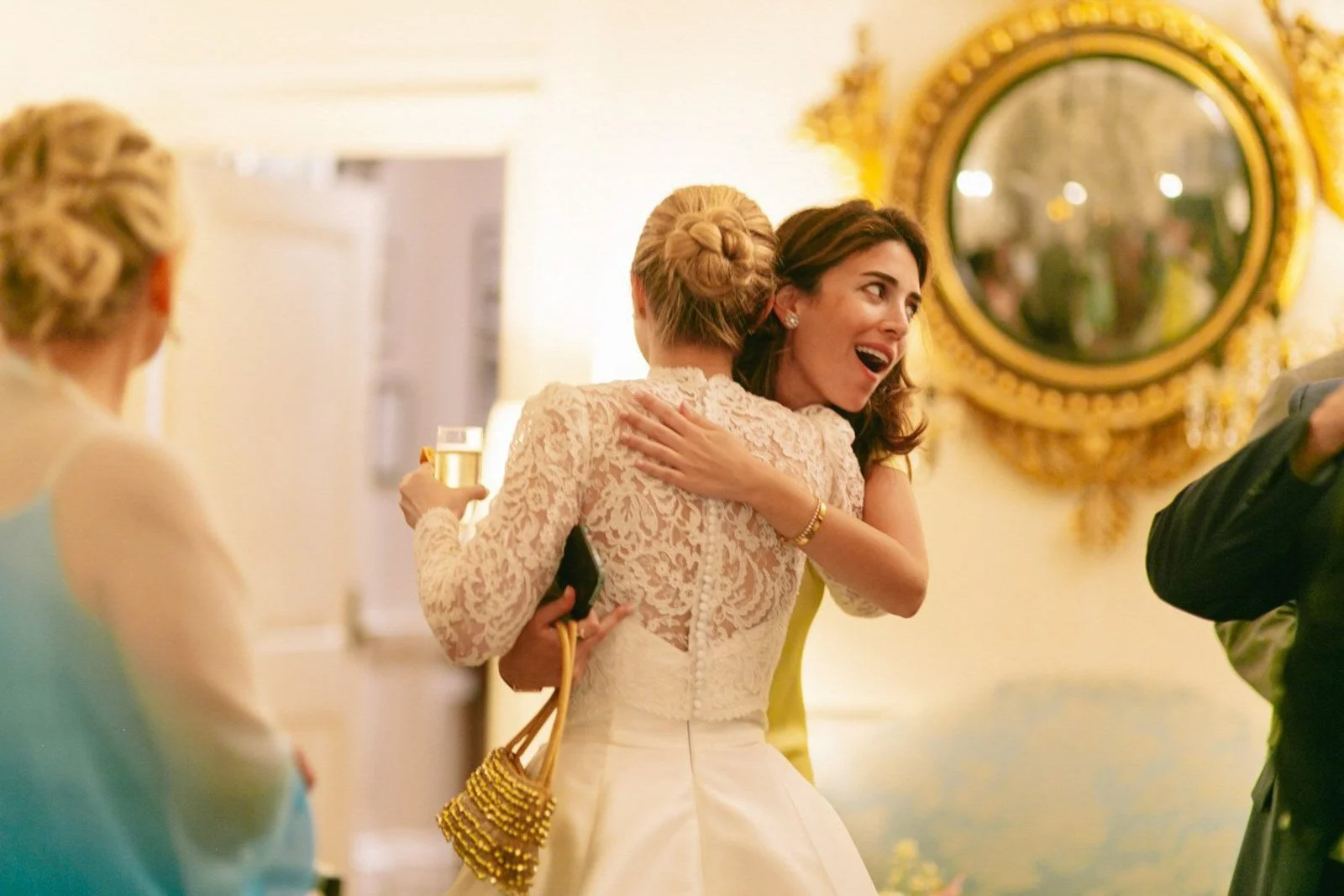 Two women embracing at a celebration, with one holding a glass of champagne. The scene is decorated with a golden mirror in the background.