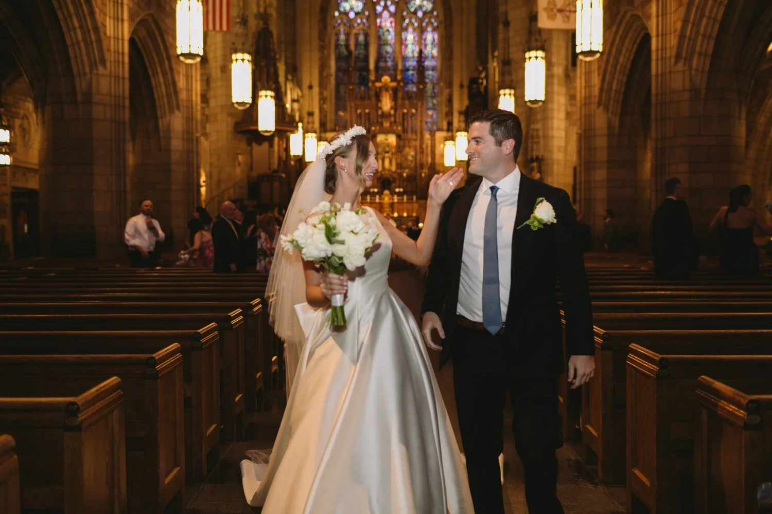 A bride and groom standing in a church, smiling at each other, with the bride holding a bouquet of white flowers.