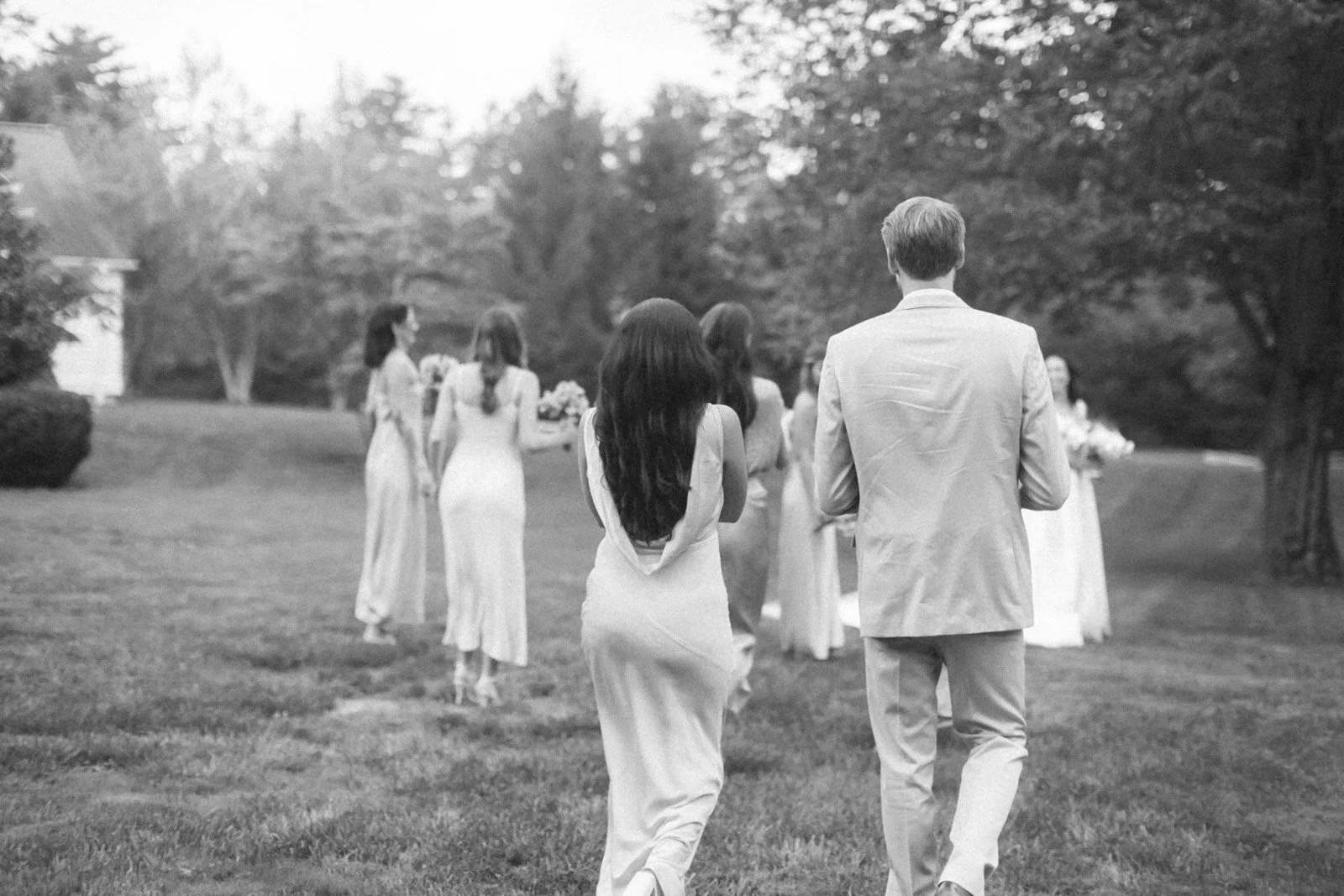 A black and white photo of a group of women and one man in elegant attire, outdoors in a park or garden setting, with trees in the background. The women are in long dresses, holding flowers, while the man wears a light-colored suit with his back to t