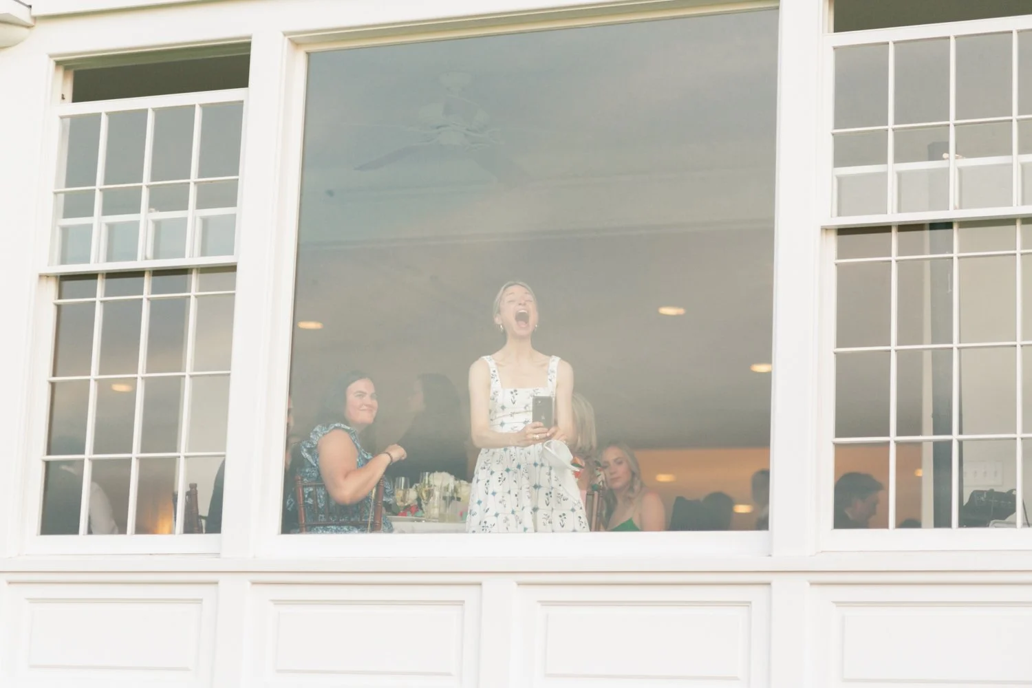 People celebrating inside a building viewed through a large window, with a woman in a white dress singing or shouting joyfully and others around her smiling and enjoying the occasion.