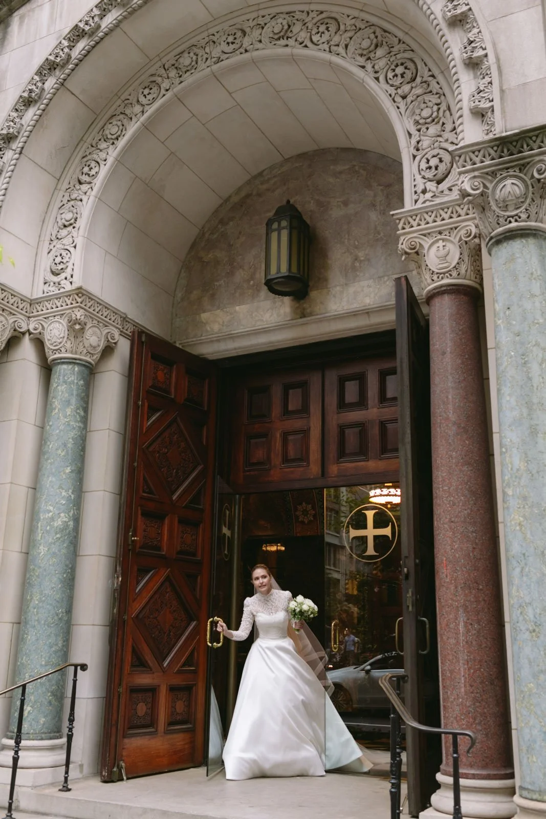 A bride in a white wedding dress and lace top, holding a bouquet, is standing at the open door of a church with ornate stone archway and columns, and a lantern hanging above.