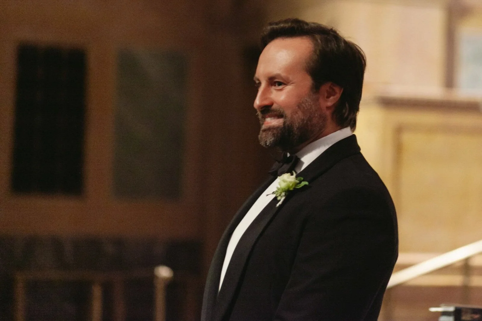 A man in a tuxedo with a boutonnière, smiling in a formal setting.