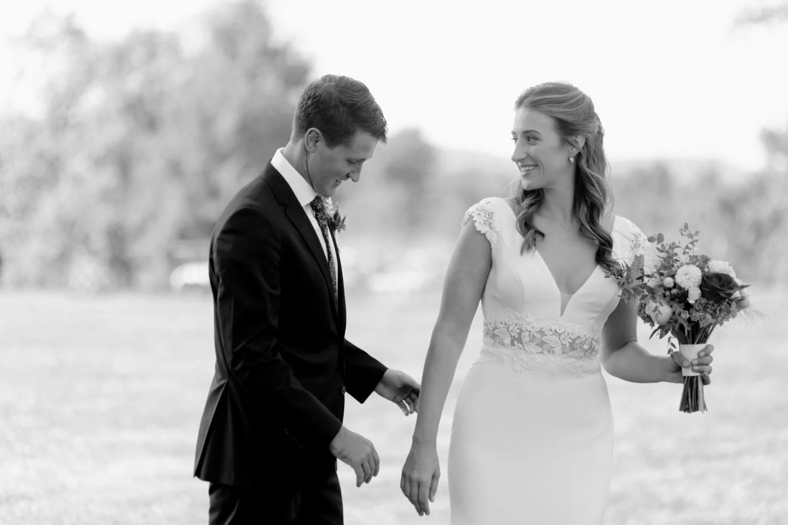 A black-and-white photo of a bride and groom outdoors, holding hands. The bride is smiling, wearing a white gown with lace details and holding a bouquet. The groom is looking down, dressed in a suit with a boutonnière, and is smiling.