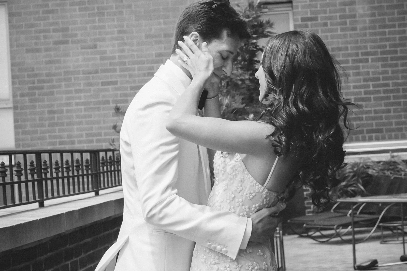 A black and white photo of a couple dancing closely. The man is in a white tuxedo and the woman is in a light-colored, sleeveless dress with floral appliqués. They are smiling and looking at each other, with the woman's hands on the man's face and th
