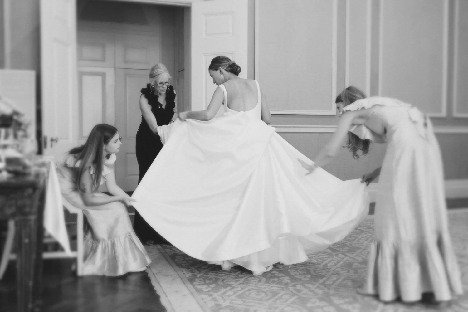 Four women helping a bride get ready, with one woman adjusting the bride's gown while two others hold the hem of the dress.