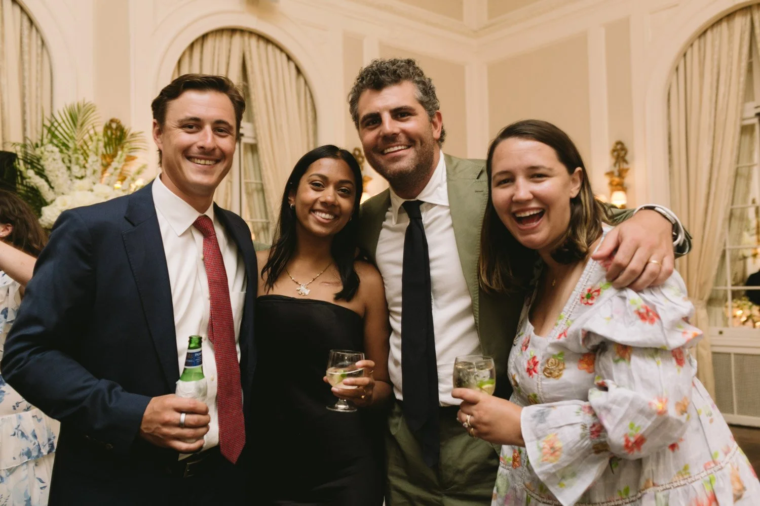Group of four friends smiling and holding drinks at a party in an elegant room with high ceilings and floral arrangements.