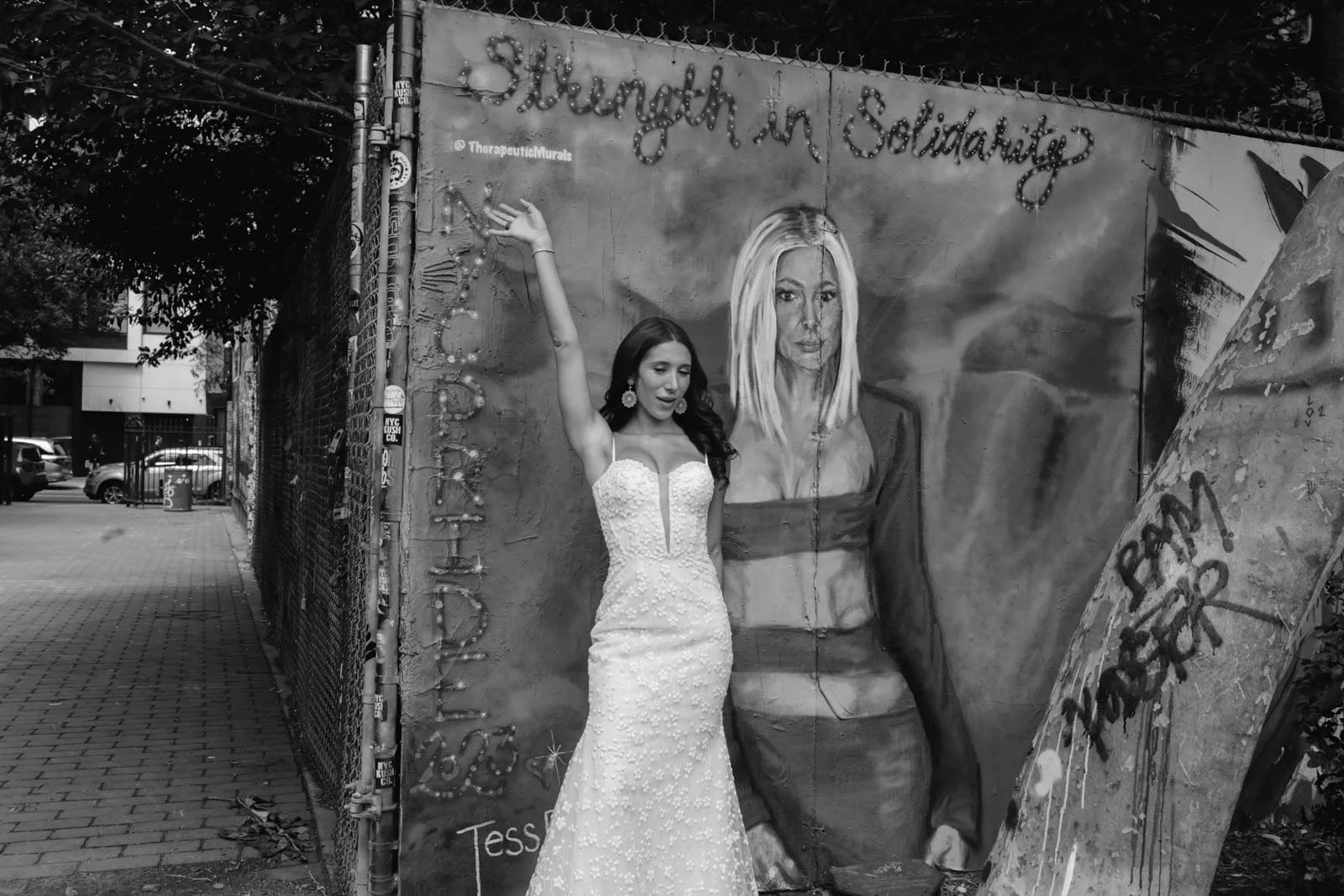 Woman in a white wedding dress posing next to a mural with a woman's portrait and the words "Strength in Solidarity" on a city sidewalk.