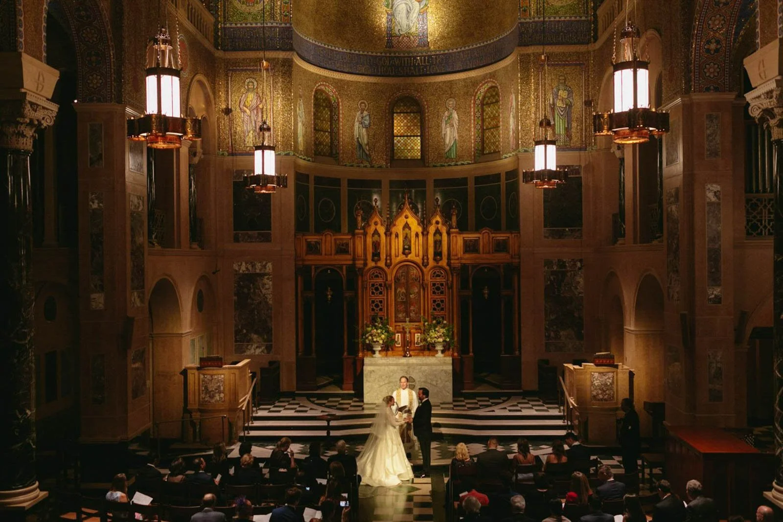 A wedding ceremony inside a church with ornate golden decorations and religious icons on the wall. The bride and groom stand at the altar with a priest officiating. Guests are seated and observing the ceremony.