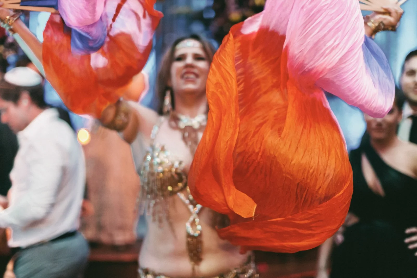 A woman in a belly dance costume with gold jewelry and a beaded top doing a dance performance at a festive event, surrounded by people in formal attire.